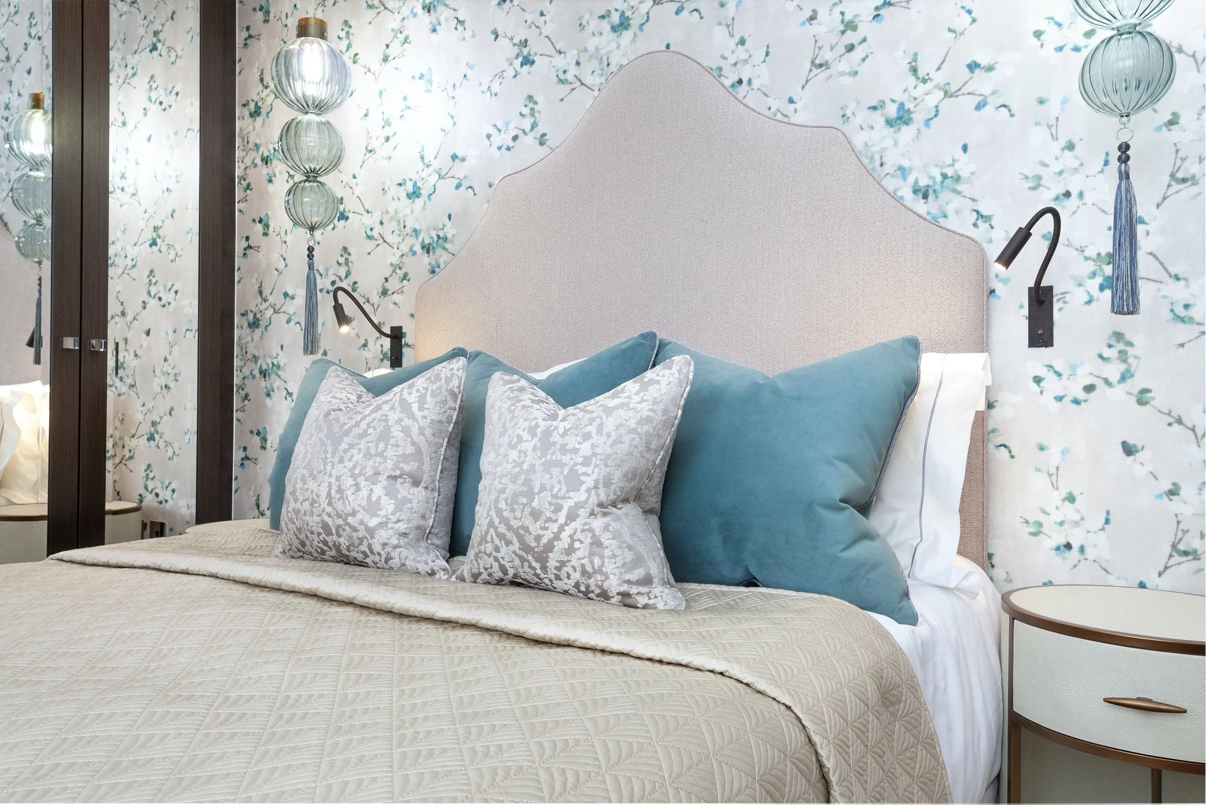 A bedroom with a bed, large pillows, a tufted headboard, decorative lamps, and patterned wallpaper featuring blue and green floral motifs.