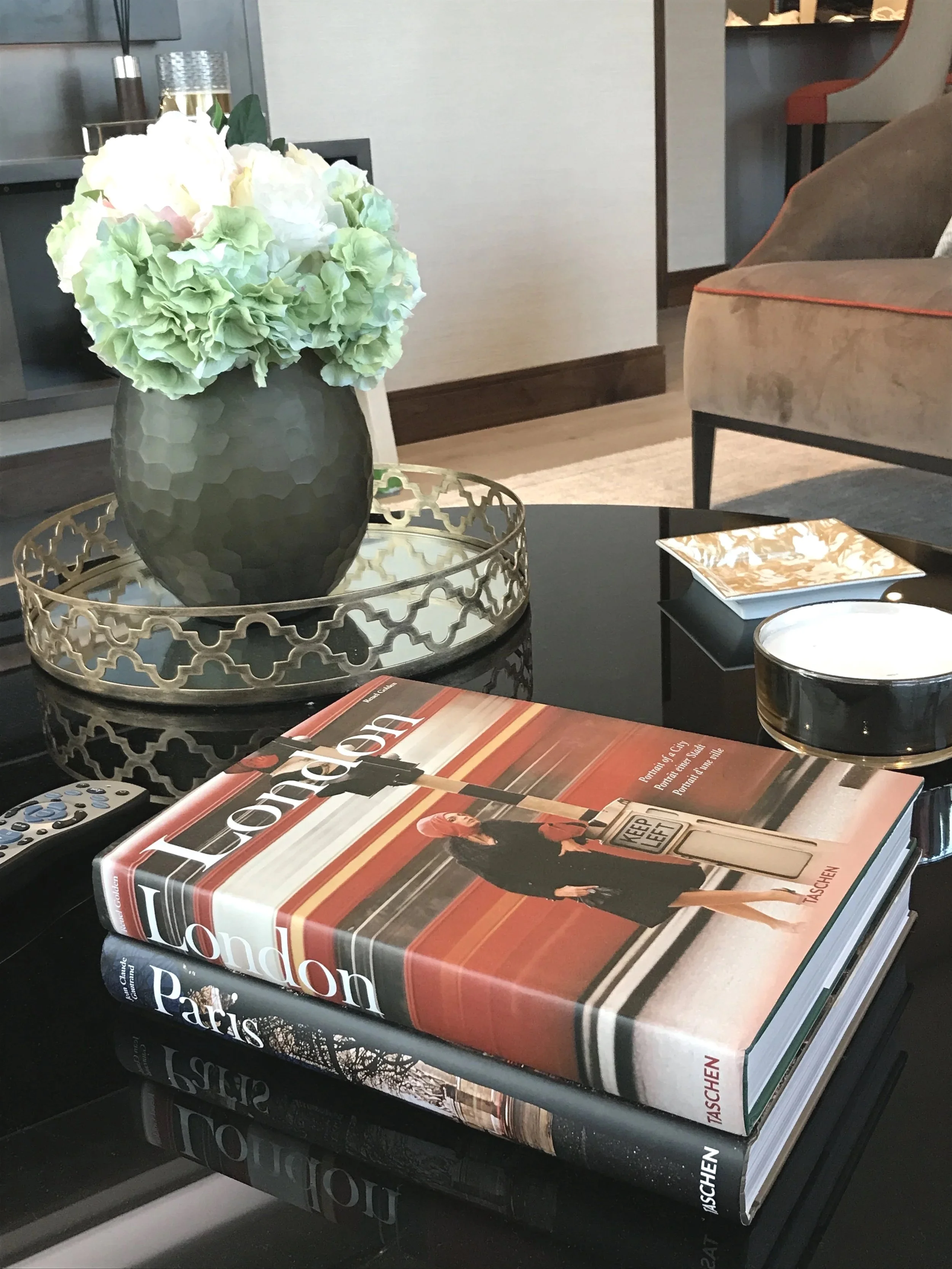 A black table with a green and white flower arrangement in a geometric vase on top, two large travel books titled "London" and "Paris," a small round black and white container, a beige coaster, a small glossy booklet, and a TV remote control. The table is in a living room with a beige chair with orange piping in the background.