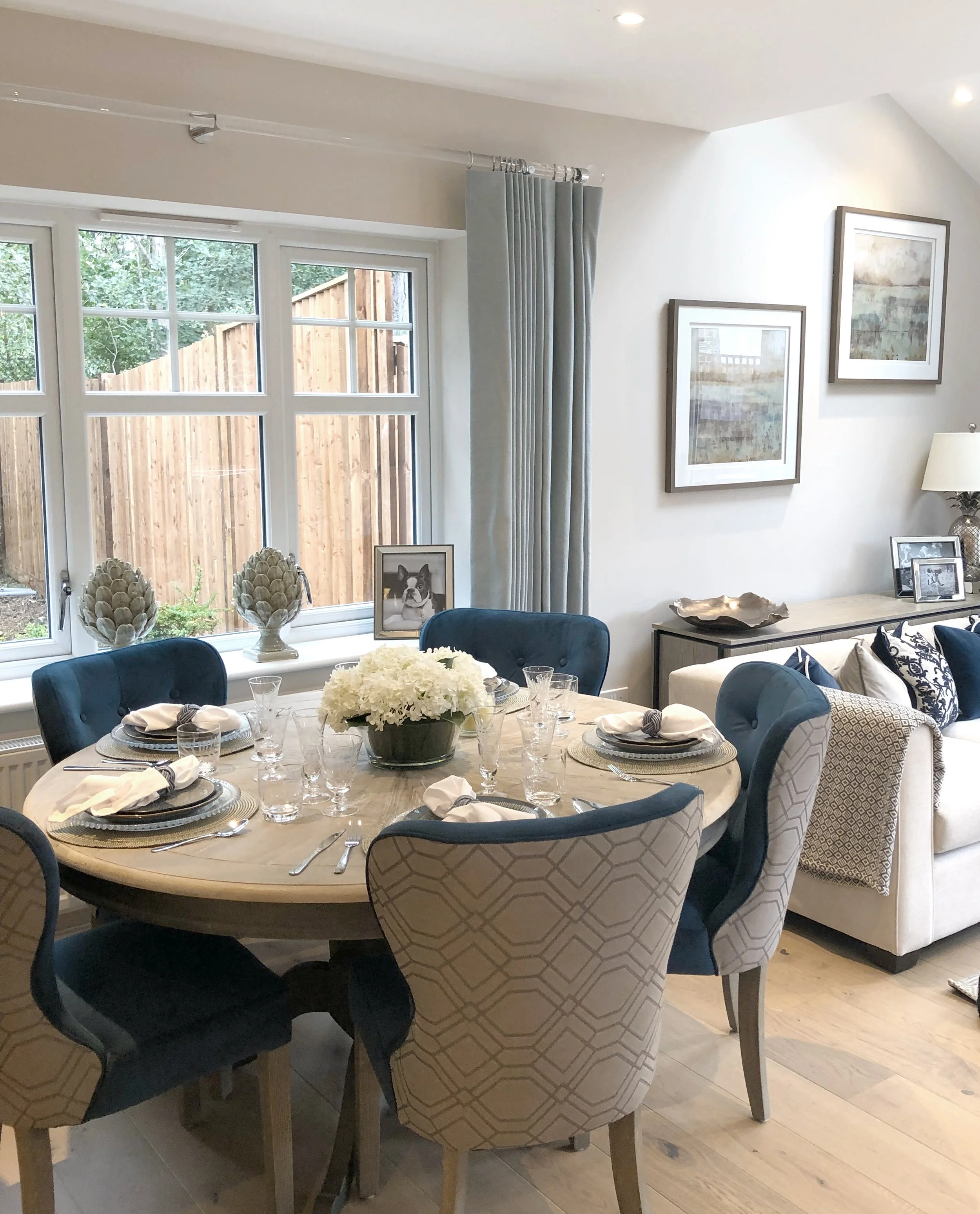 A dining room with a round table set for a meal. The table has a floral centerpiece, placemats, napkins, glasses, and silverware. There are six upholstered chairs around the table, with some in dark blue and some in a light patterned fabric. The room has a large window with light blue curtains, framed artwork on the walls, and a white sofa with decorative pillows in the background.