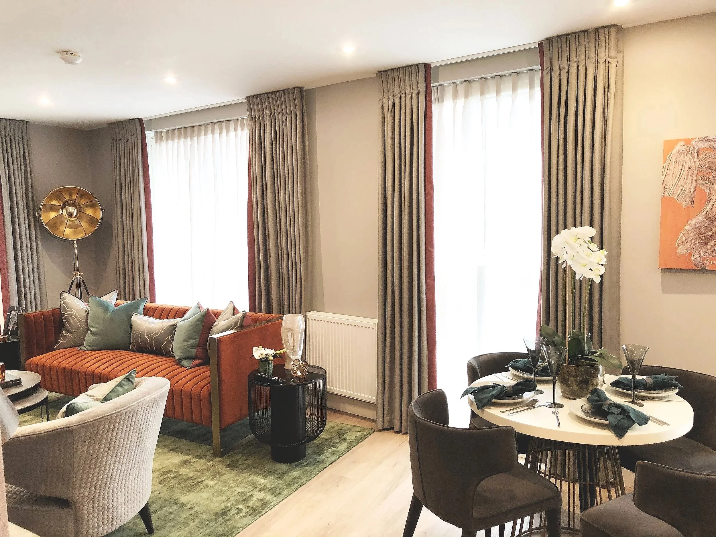 A living room and dining area with large windows, beige walls, beige curtains, a striped orange sofa with pillows, a white armchair, a round dining table with black chairs, tableware, napkins, a centerpiece with white orchids, artwork on the wall, and a floor lamp.