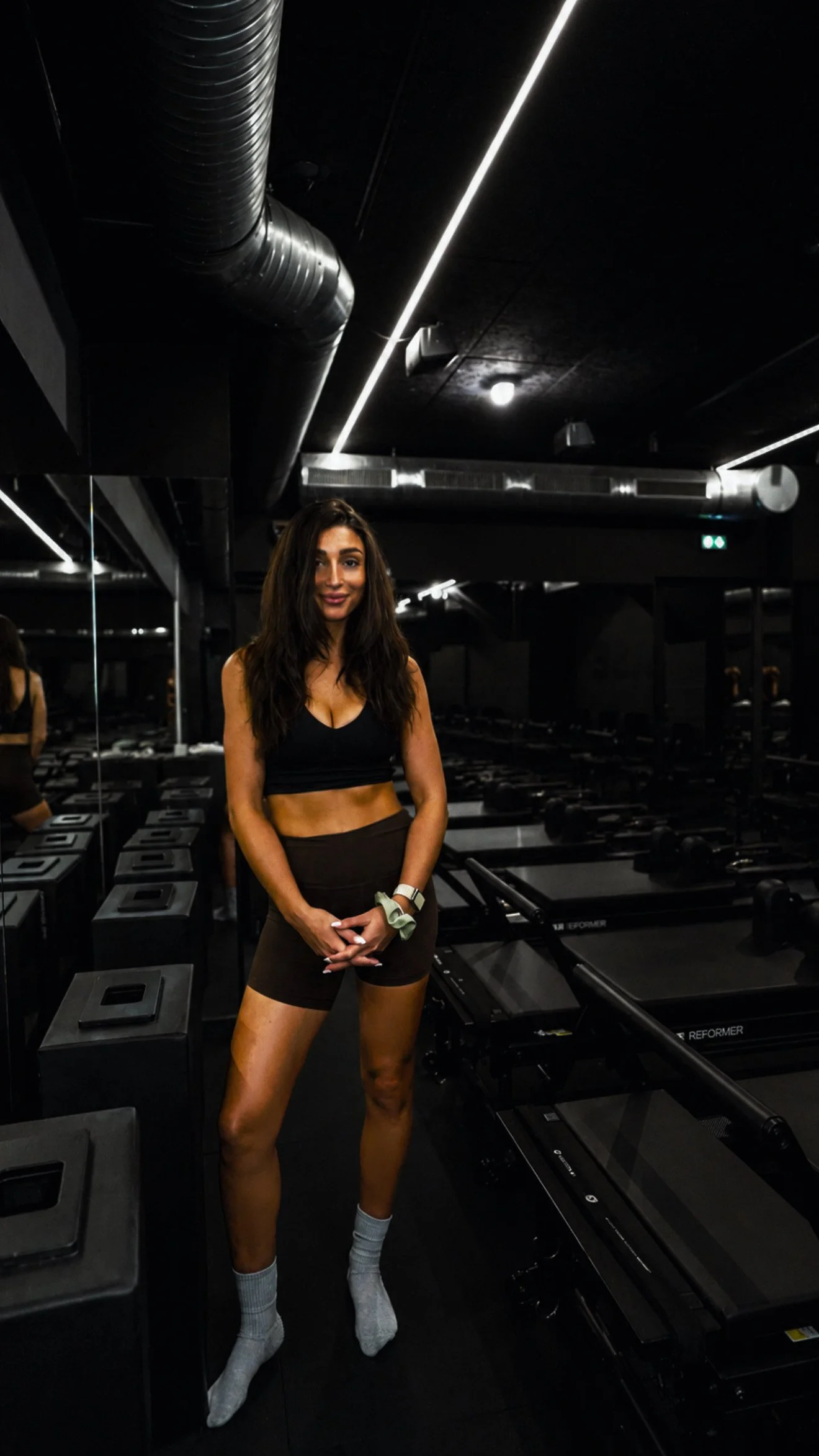A woman in athletic workout attire standing in a gym, with black workout equipment and mirrors in the background.
