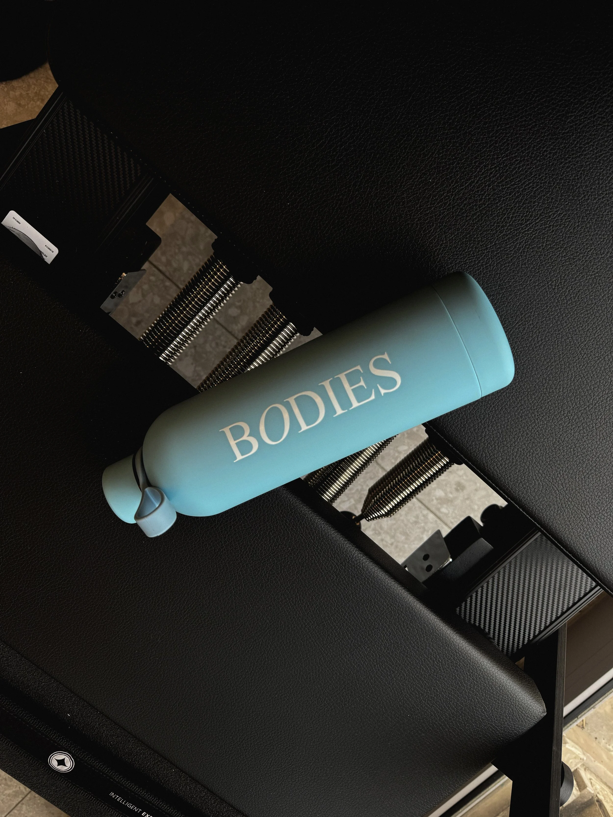 A light blue Bodies water bottle with a white cap placed on a black textured surface, next to a mechanical device with springs.