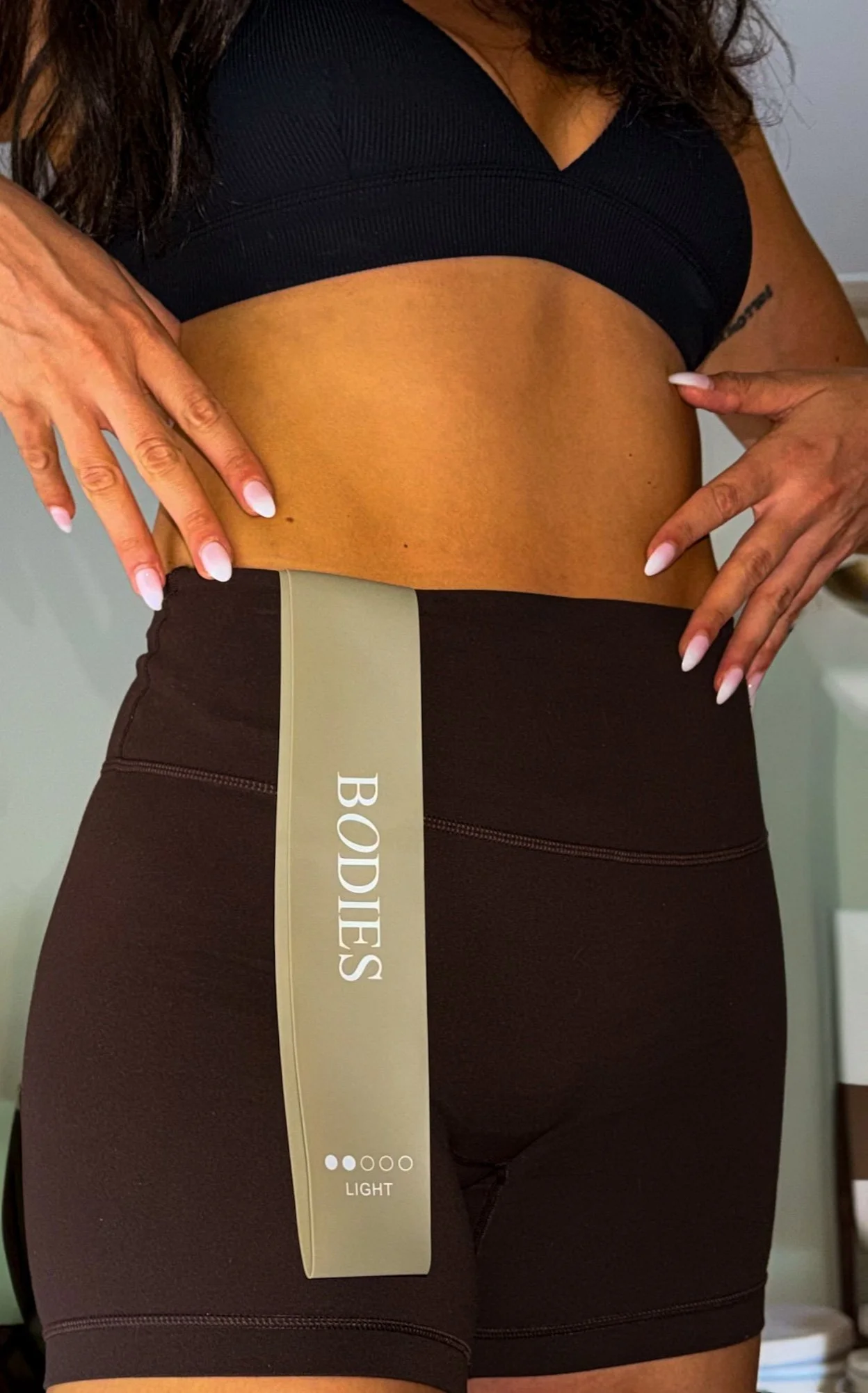Close-up of a woman's midsection wearing black tights with a beige waistband. The woman is holding her waist with both hands. The tights band has the text 'BODIES' on it and indicates a 'LIGHT' level.