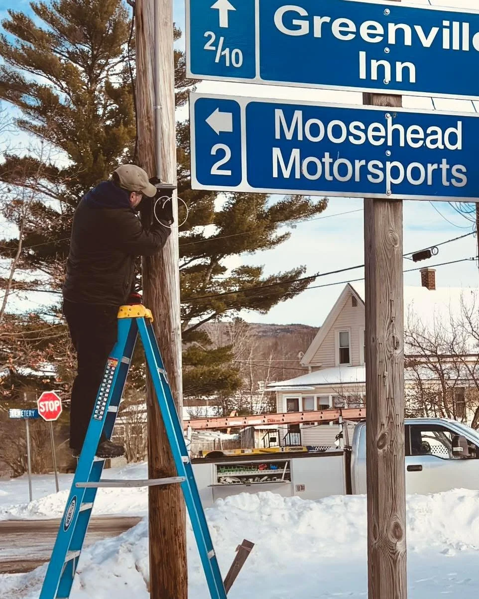 A man on a ladder working on a wooden utility pole next to blue road signs for Greenville Inn and Moosehead Motorsports in a snowy outdoor setting.