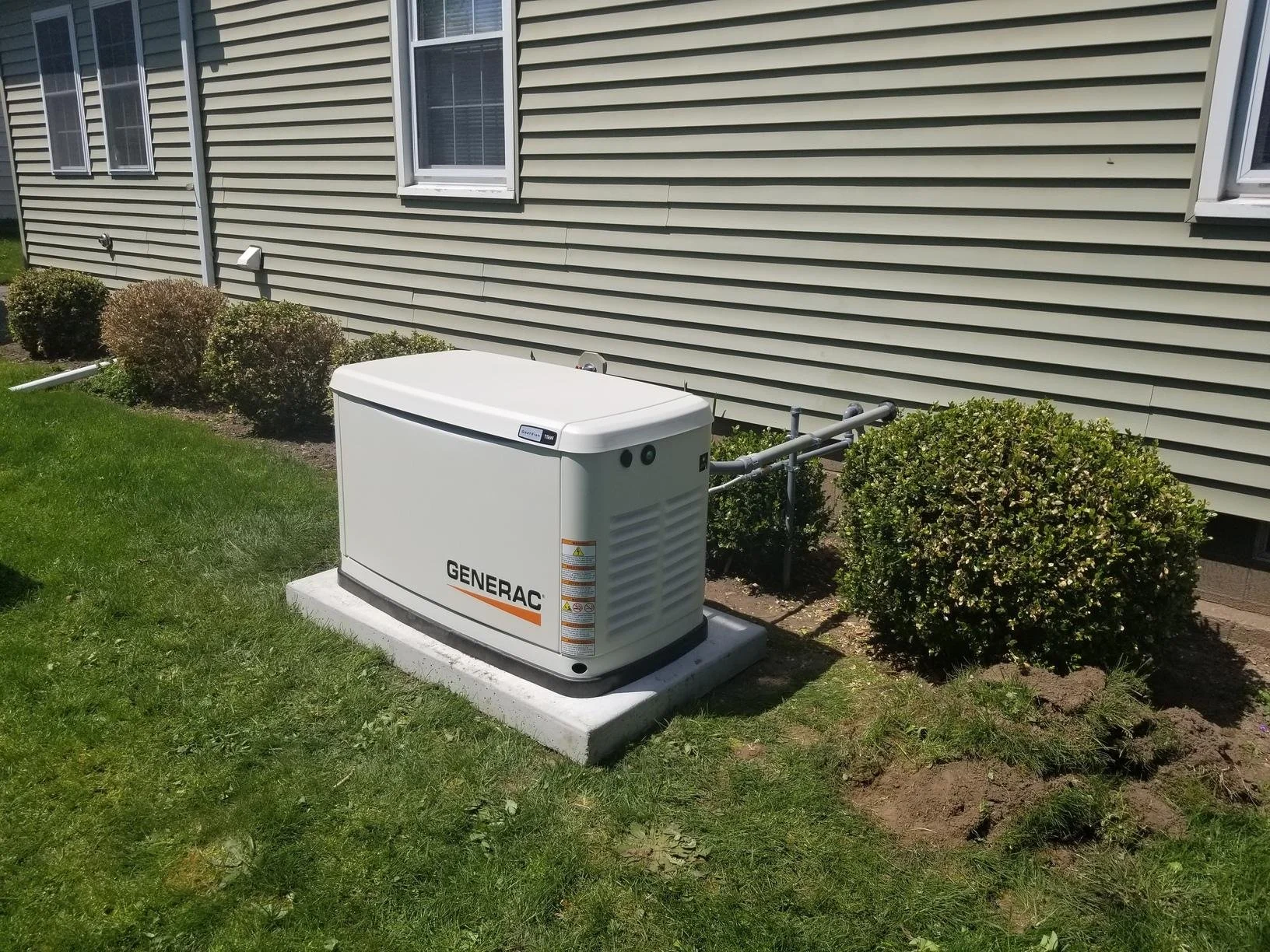Generac backup generator installed outside the beige house with green siding, next to small neatly trimmed bushes and a grassy lawn.