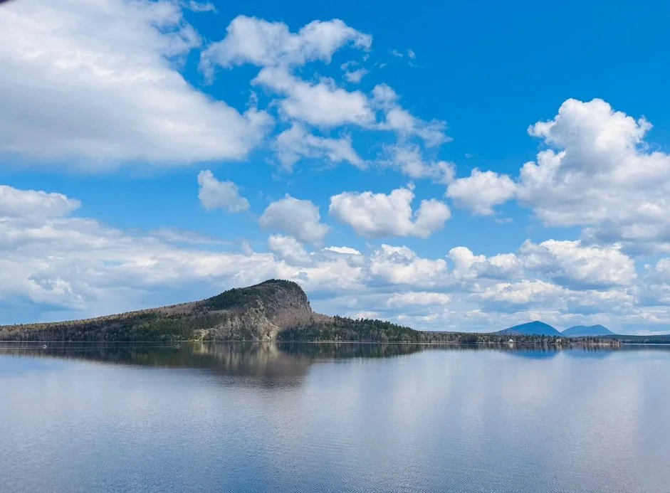 Scenic view of a large lake with a hilly landscape in the background, under a partly cloudy blue sky.