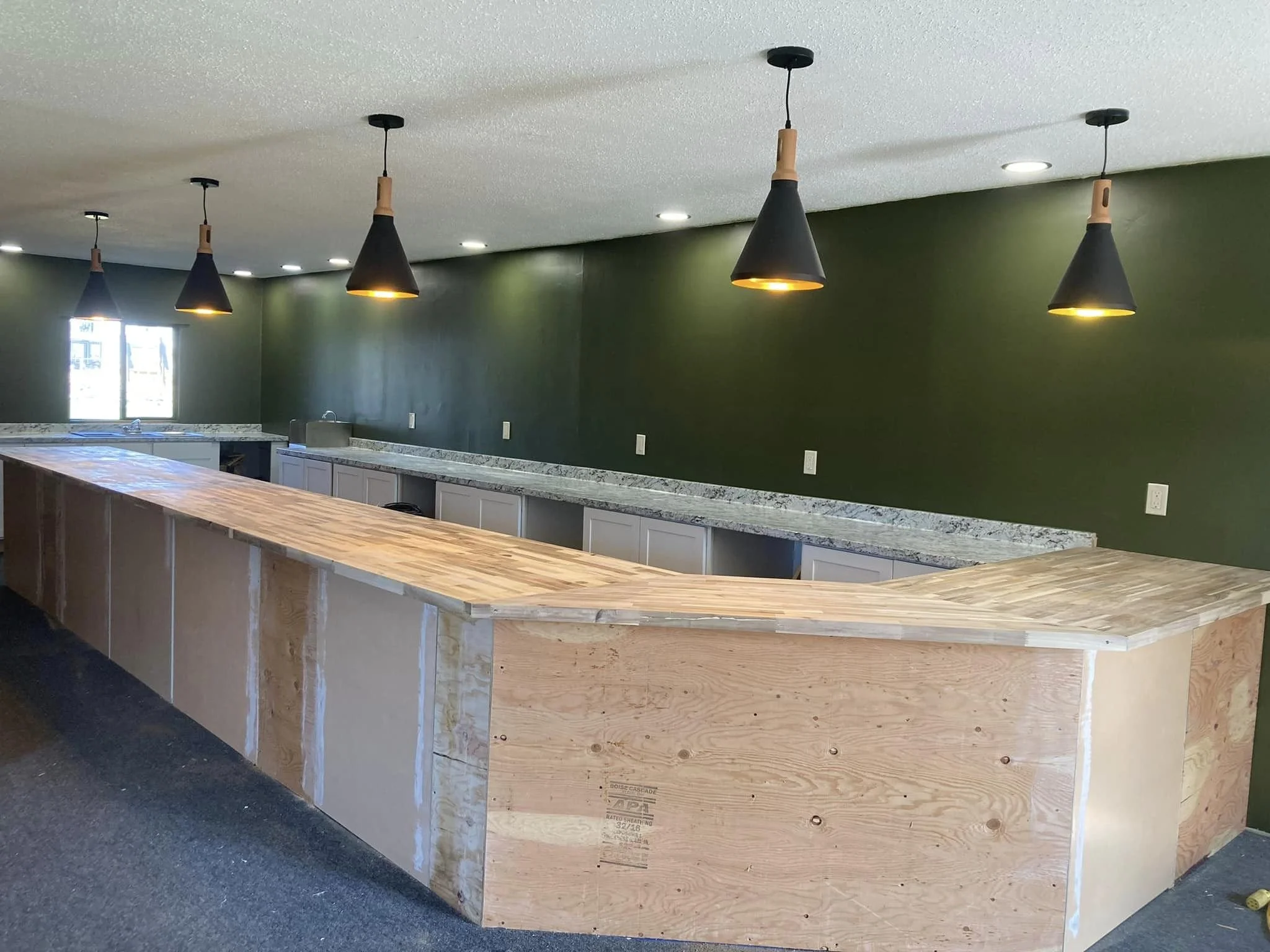 Bar area under construction with a wooden countertop, green wall, pendant lights, and a window in the background.