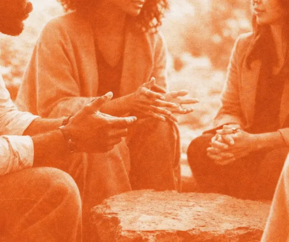 Four individuals sitting in a circle, engaged in a conversation, with one person's hands gesturing. The setting appears to be outdoors, with a rocky surface in the foreground.