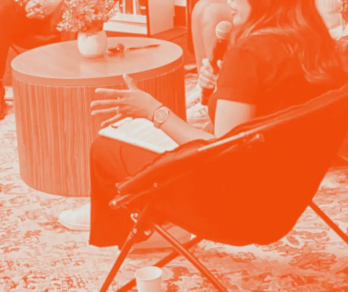 Person sitting in a modern indoor setting, holding a microphone, with a notebook on their lap, sitting on a foldable chair next to a round wooden table with a flower vase, in a group discussion or interview setting.
