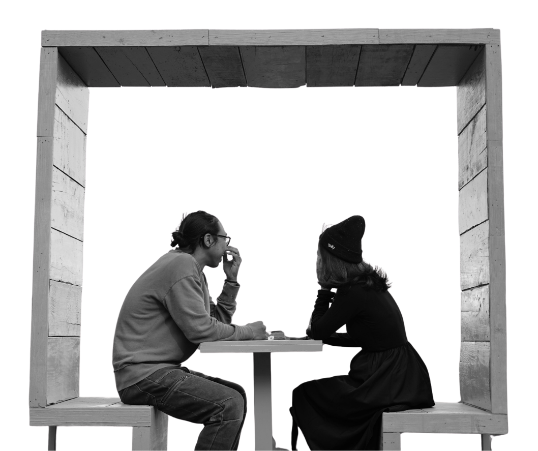 Two people sitting across from each other at a small table in a wooden booth, engaged in conversation. There is a modern, wooden cube that makes up the benches they sit in.