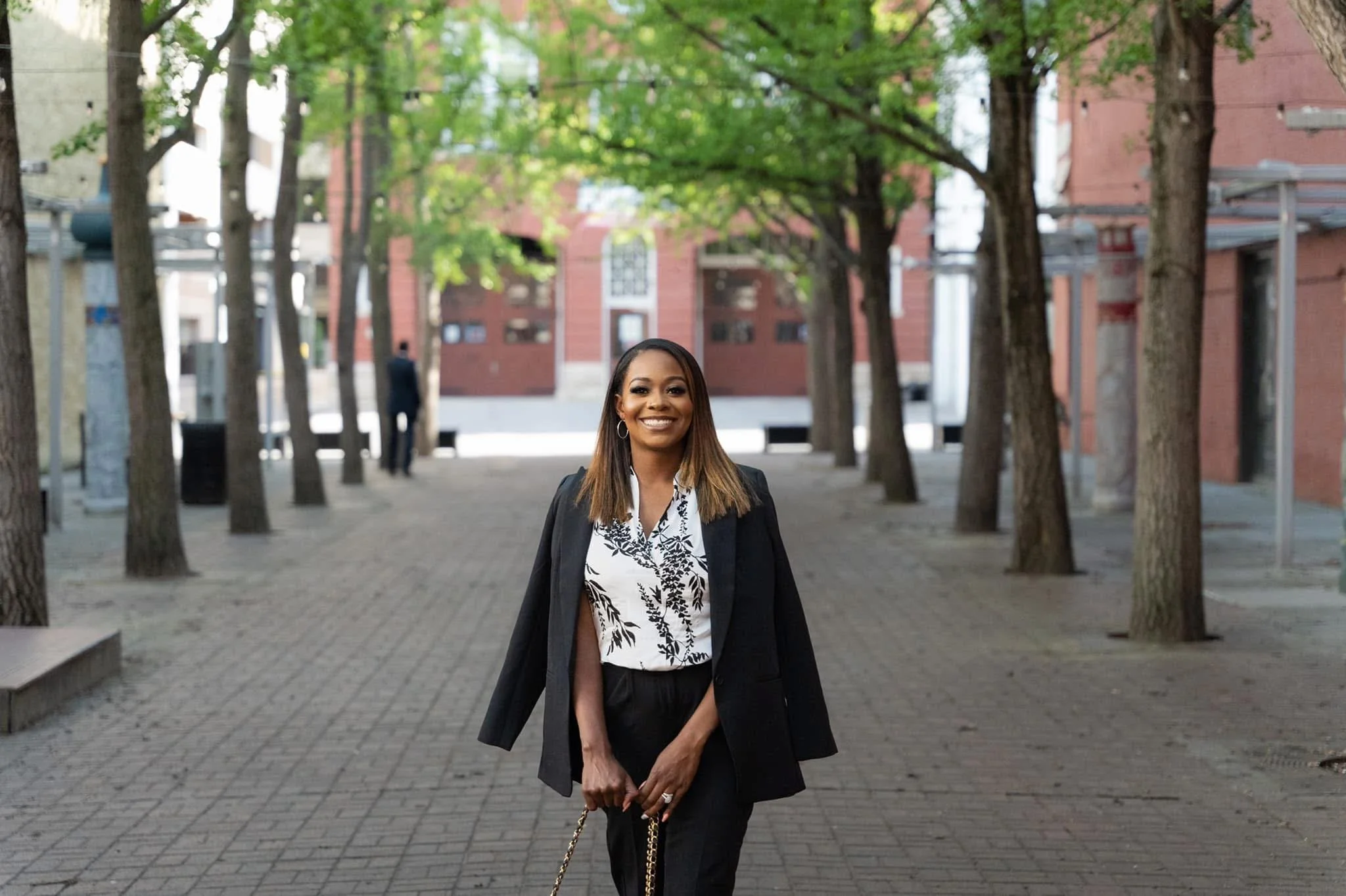 A smiling woman in business attire standing on a tree-lined brick pathway in an urban setting, with red brick buildings and a man in the background.