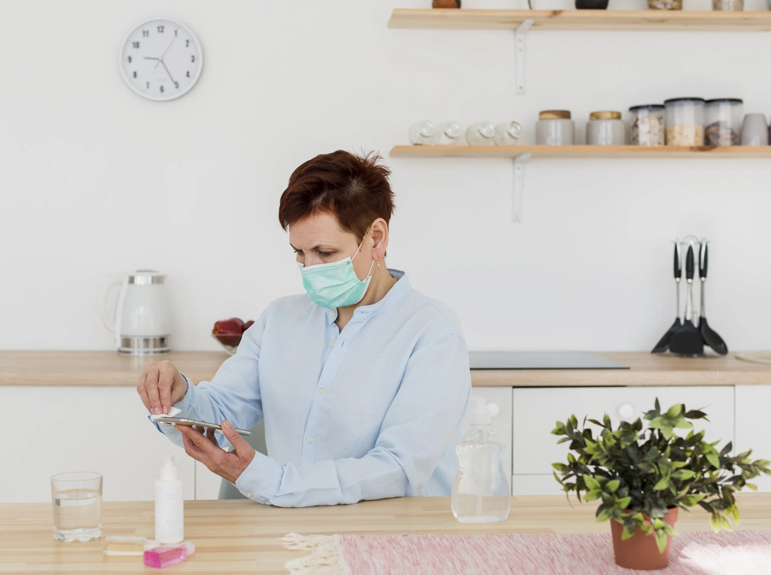 A woman with short red hair wearing a medical mask and a light blue shirt, looking at her smartphone while standing at a kitchen counter. The kitchen has white cabinets, wooden shelves with jars and containers, a clock on the wall, a glass of water, hand sanitizer, and a potted plant.