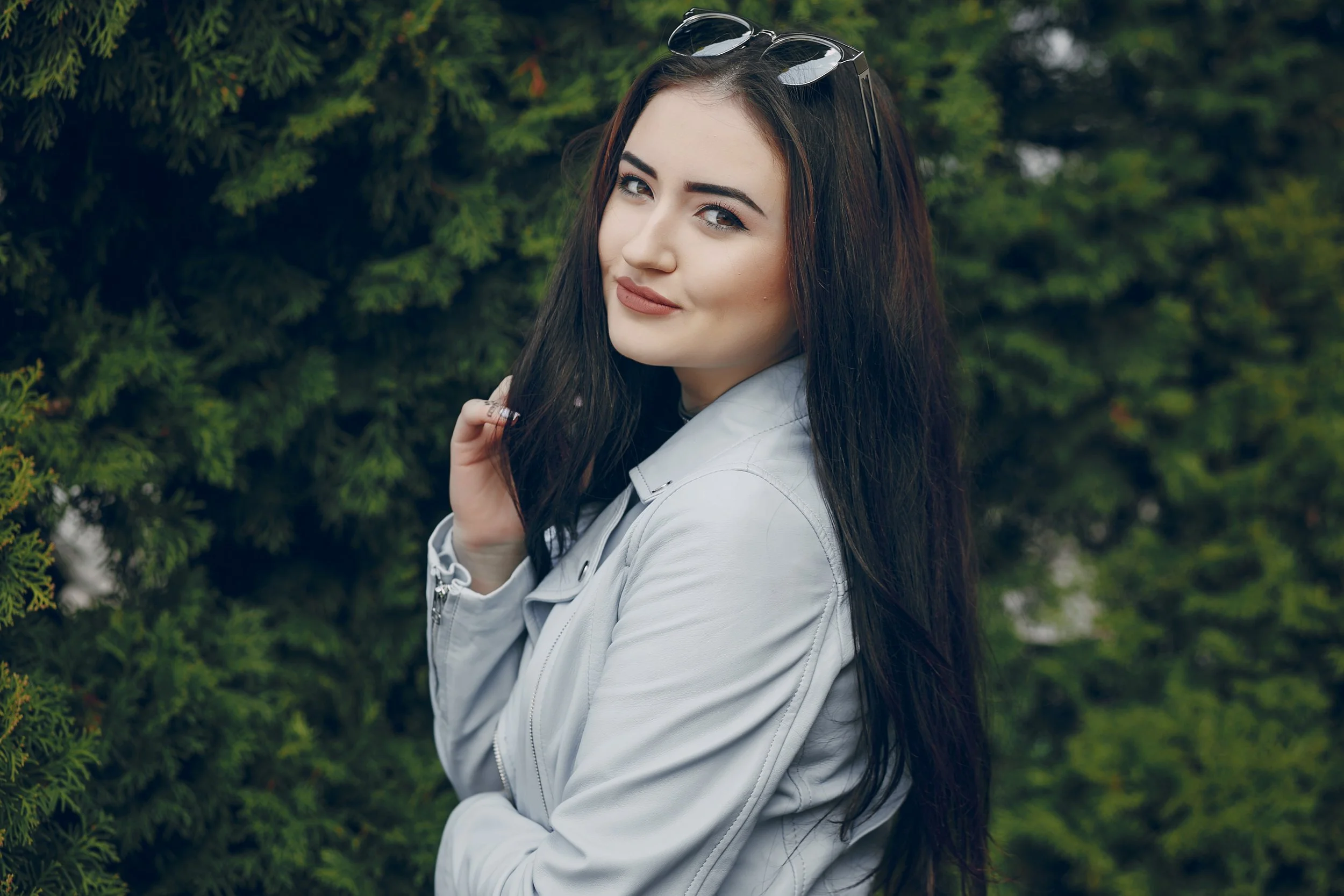 A young woman with long dark hair, wearing a light gray jacket and sunglasses on her head, posing outdoors in front of green foliage.