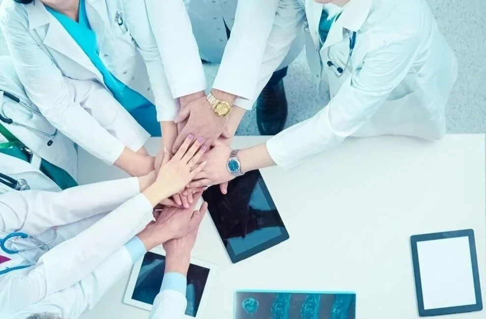 Group of healthcare professionals in white coats placing their hands together in a team gesture over a hospital table with medical tablets and X-ray images.