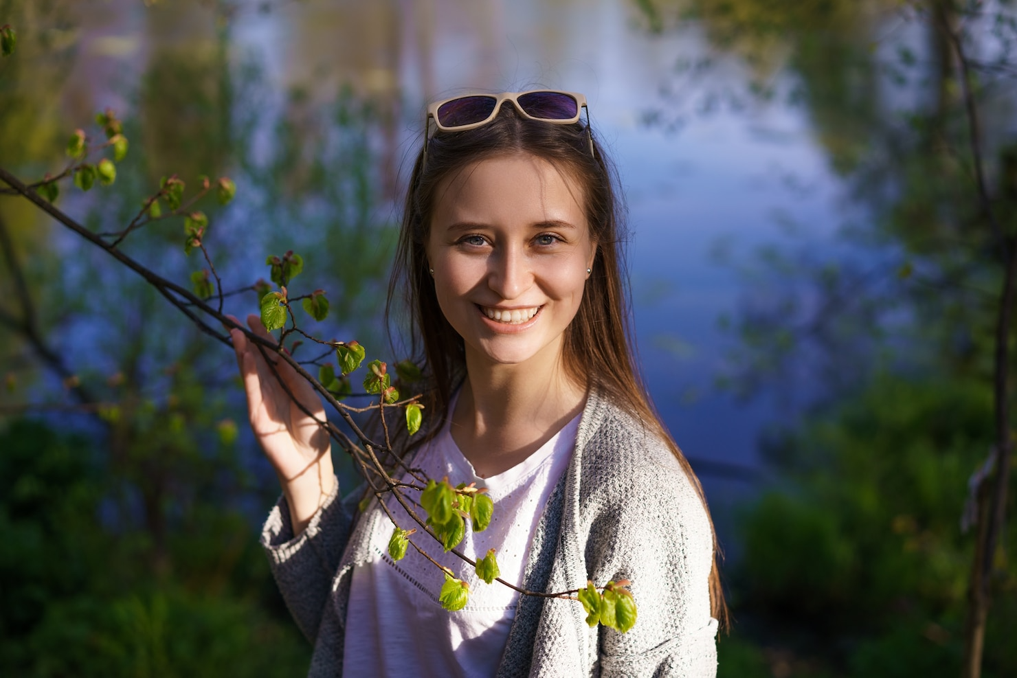 A woman with sunglasses on her head smiling outdoors near a body of water surrounded by trees with budding leaves.