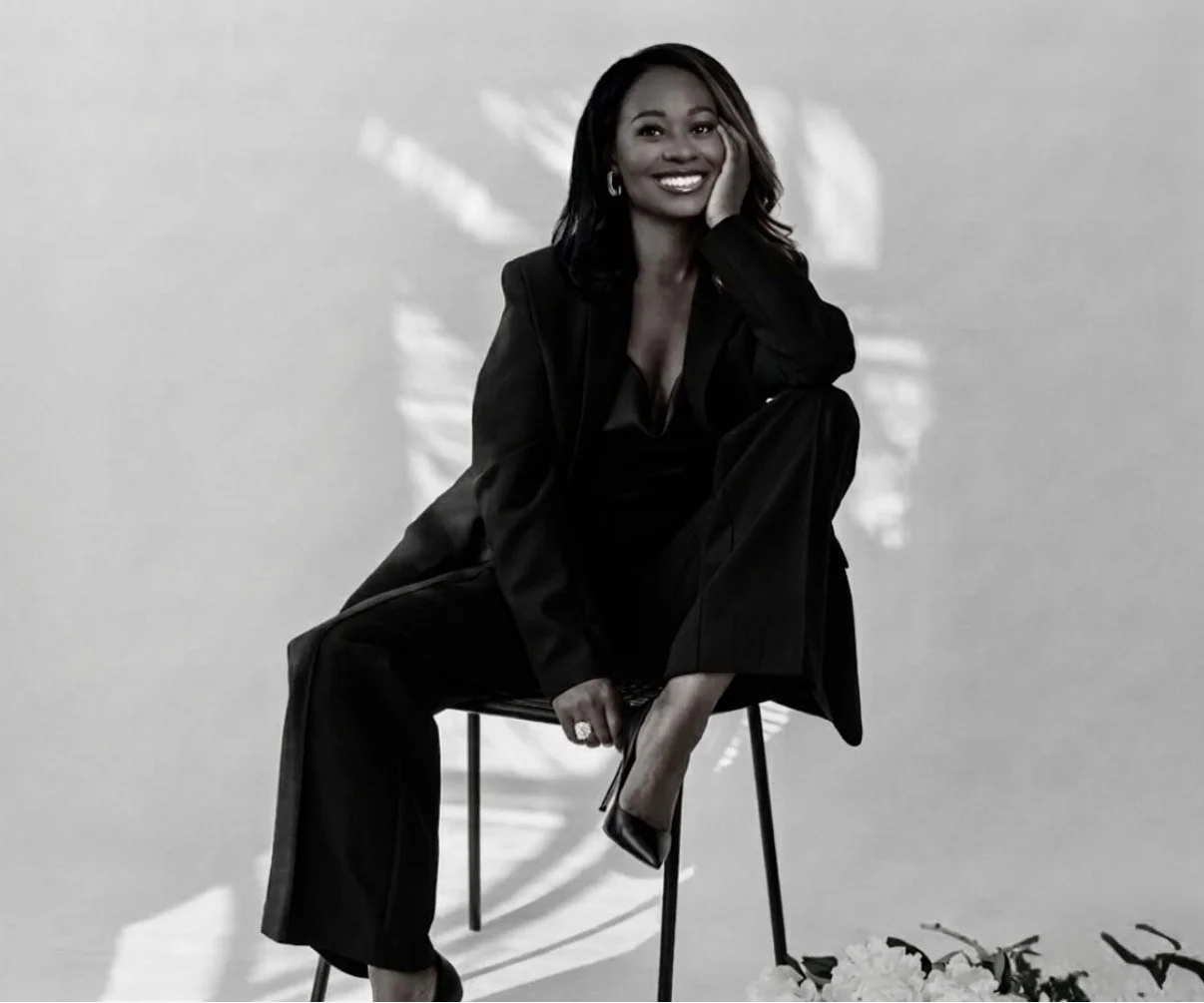 A woman dressed in black sitting on a chair, smiling, with one hand resting on her face and her other leg crossed over her knee, in front of a plain wall