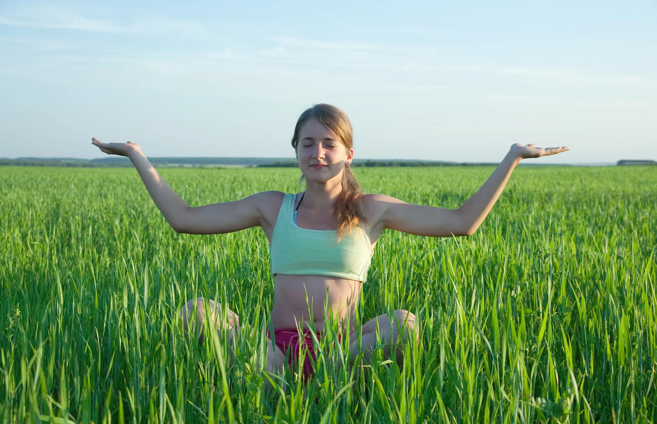 A young woman in yoga pose in a green field with her arms extended and eyes closed.