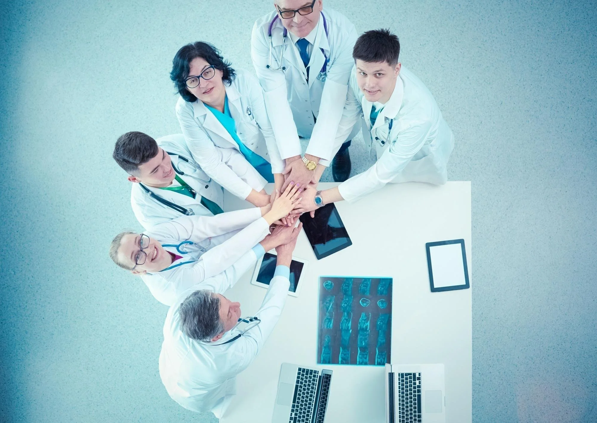 Medical team of seven doctors and nurses putting hands together in a huddle, viewed from above, with medical equipment and tablets on a white table.