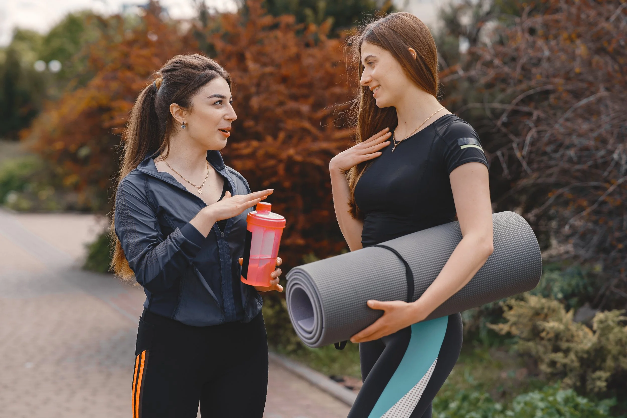Two women are having a conversation outdoors during autumn, one holding a pink water bottle and wearing a navy jacket, the other with a yoga mat, dressed in athletic wear, smiling at each other.