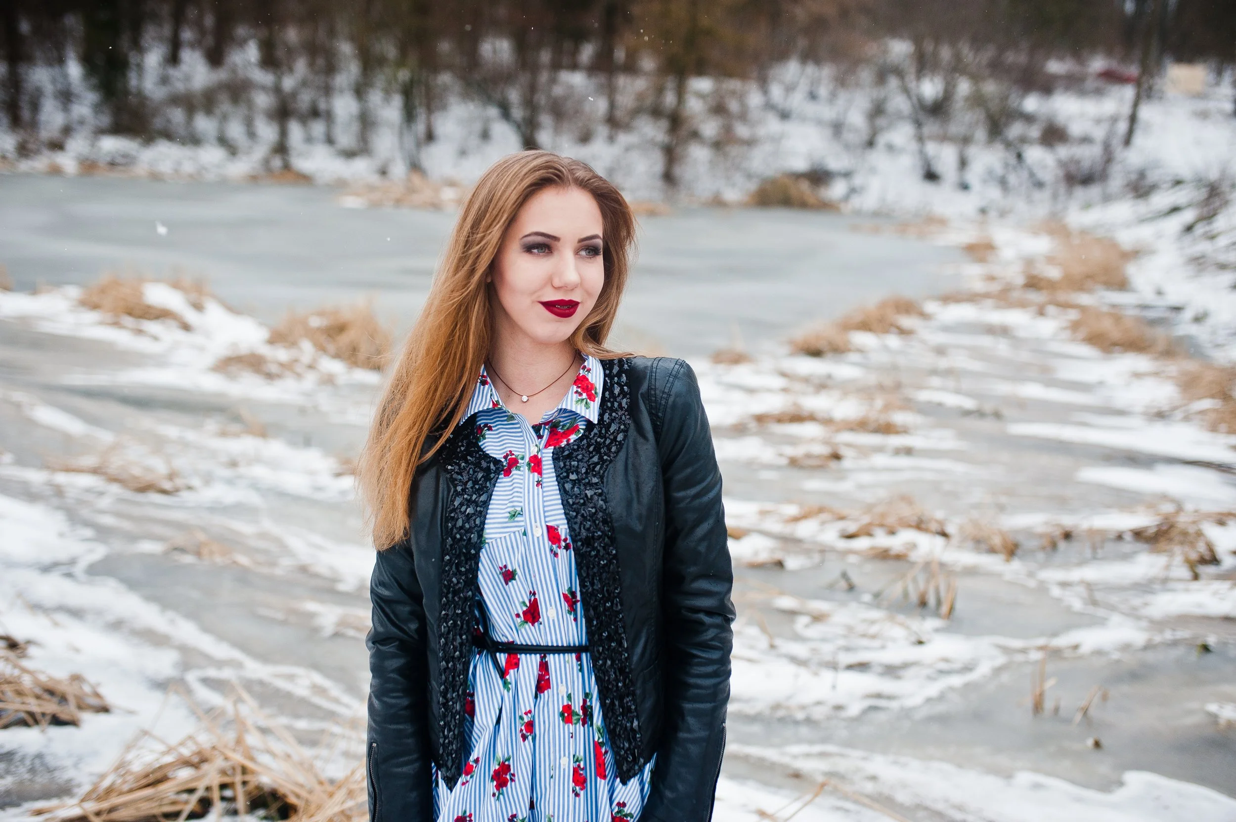 A woman with long red hair and red lipstick standing outdoors near snow-covered ground and dead plants, wearing a blue and white striped dress with red flowers and a black jacket.
