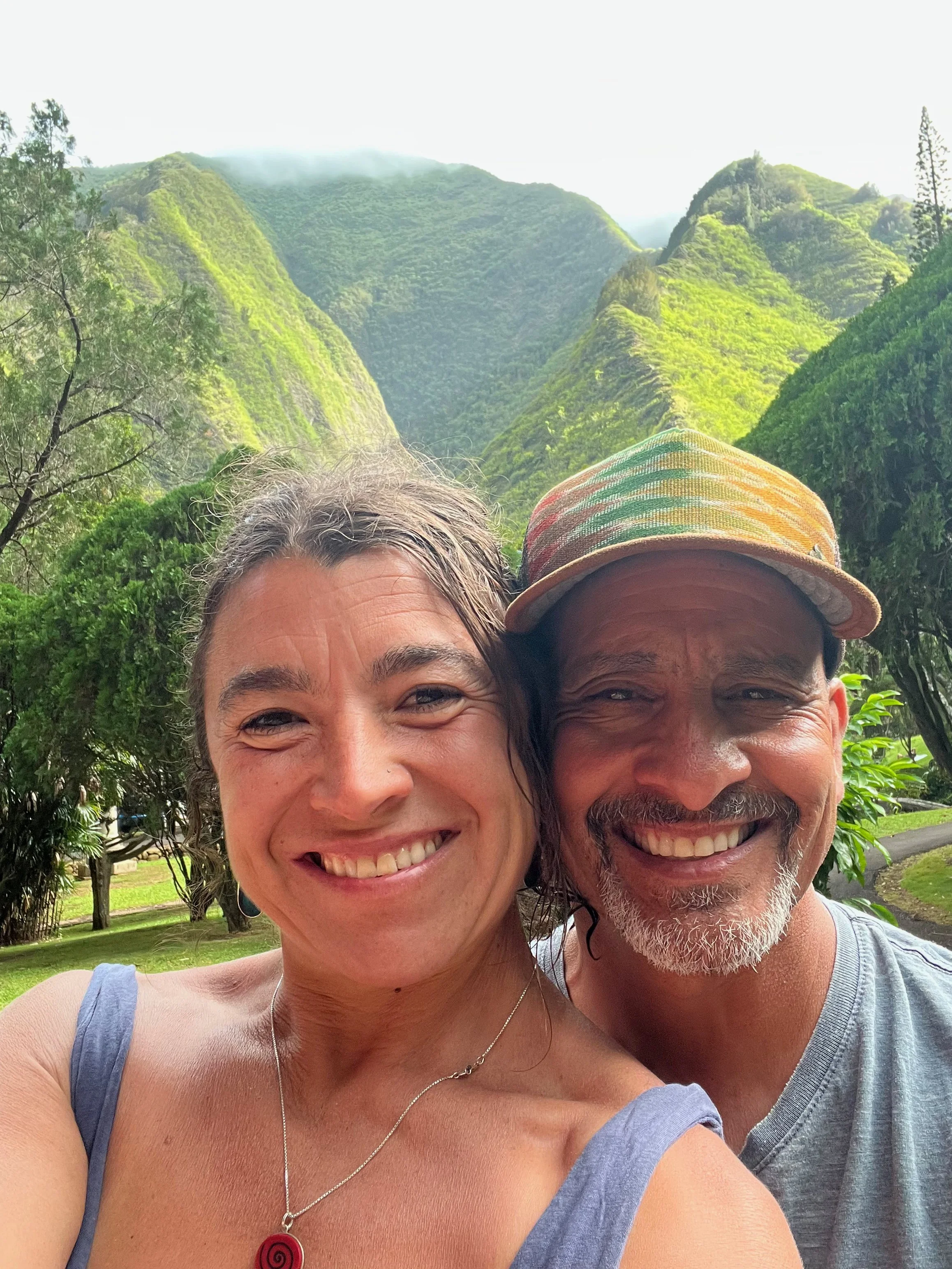 A smiling woman and man taking a selfie in front of lush green mountains.