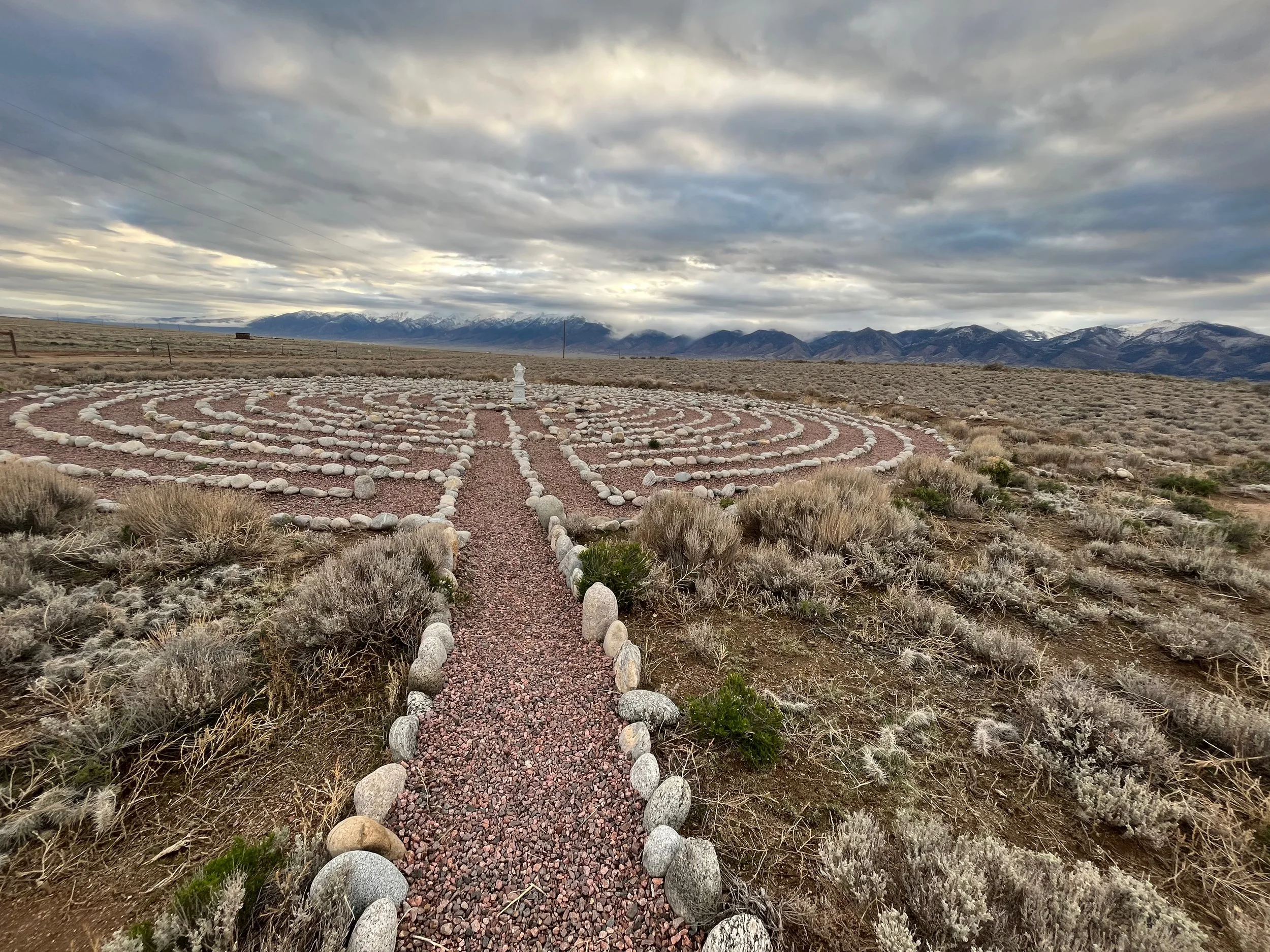Desert landscape with a labyrinth made of rocks in the foreground, a mountain range in the background, and a cloudy sky overhead.