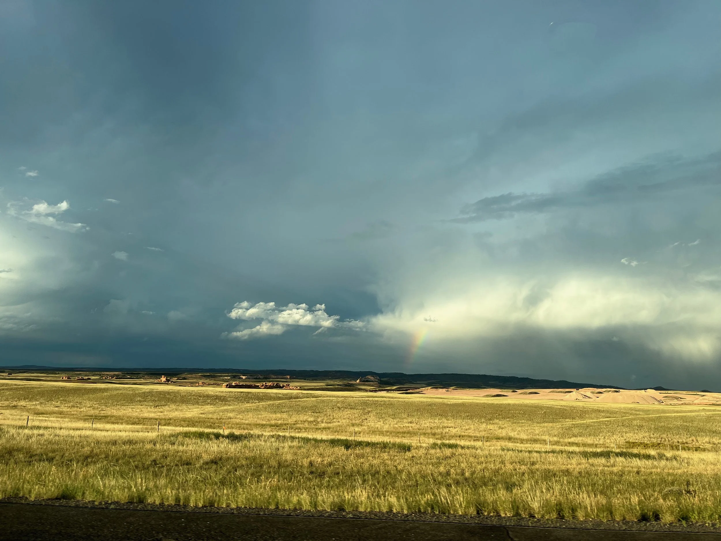 A wide open field with green grass and small structures in the distance, under a dramatic sky with dark clouds and a faint rainbow.
