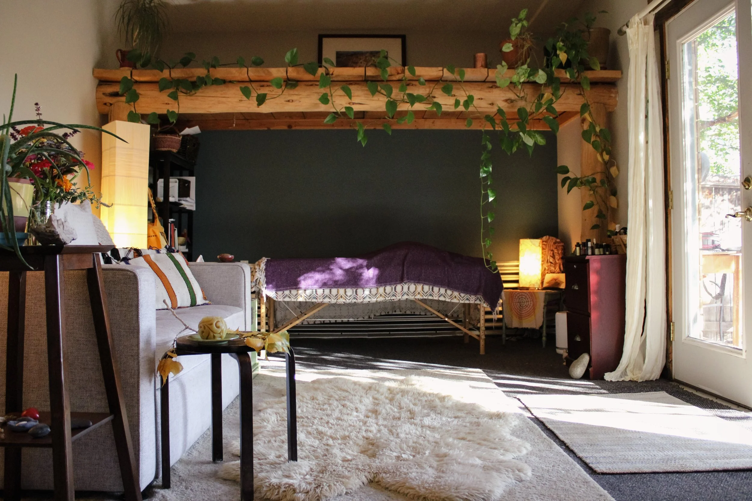 Living room with a beige sofa, small black coffee table, fluffy white rug, houseplants, a cozy bed, and a glass door leading outside, with sunlight streaming in.
