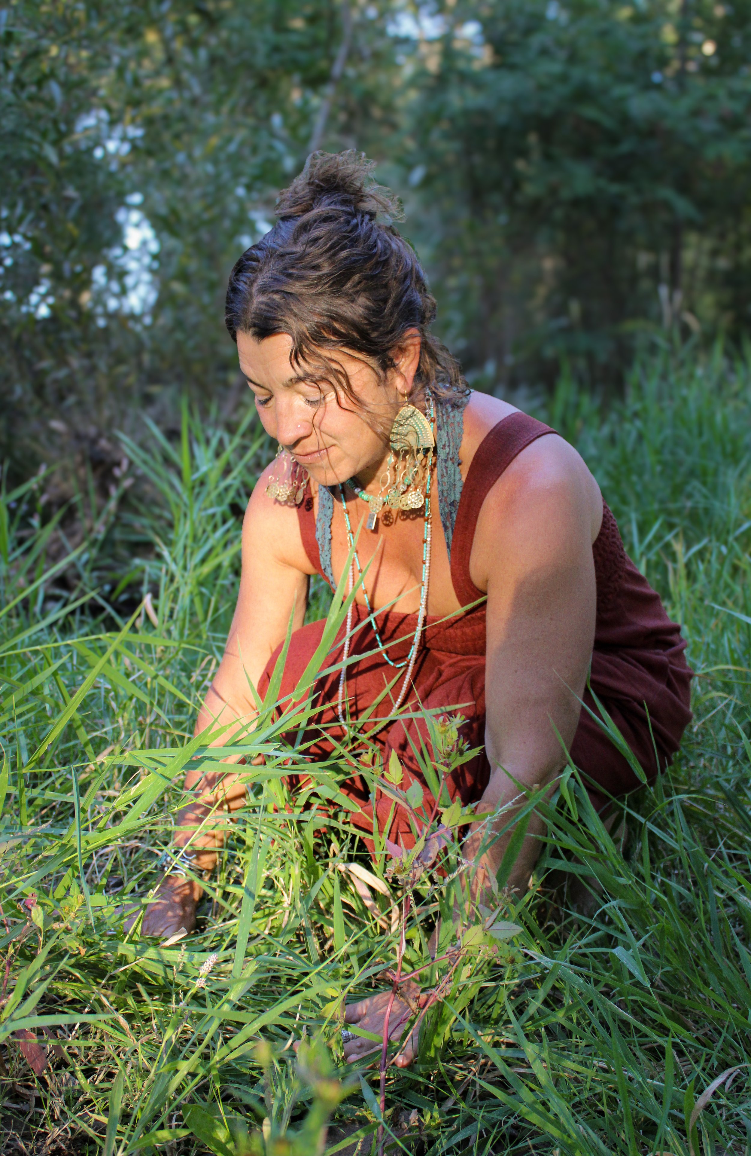 A woman with dark, curly hair in a bun, wearing bohemian jewelry, crouching in tall grass outdoors, looking down and tending to the plants.