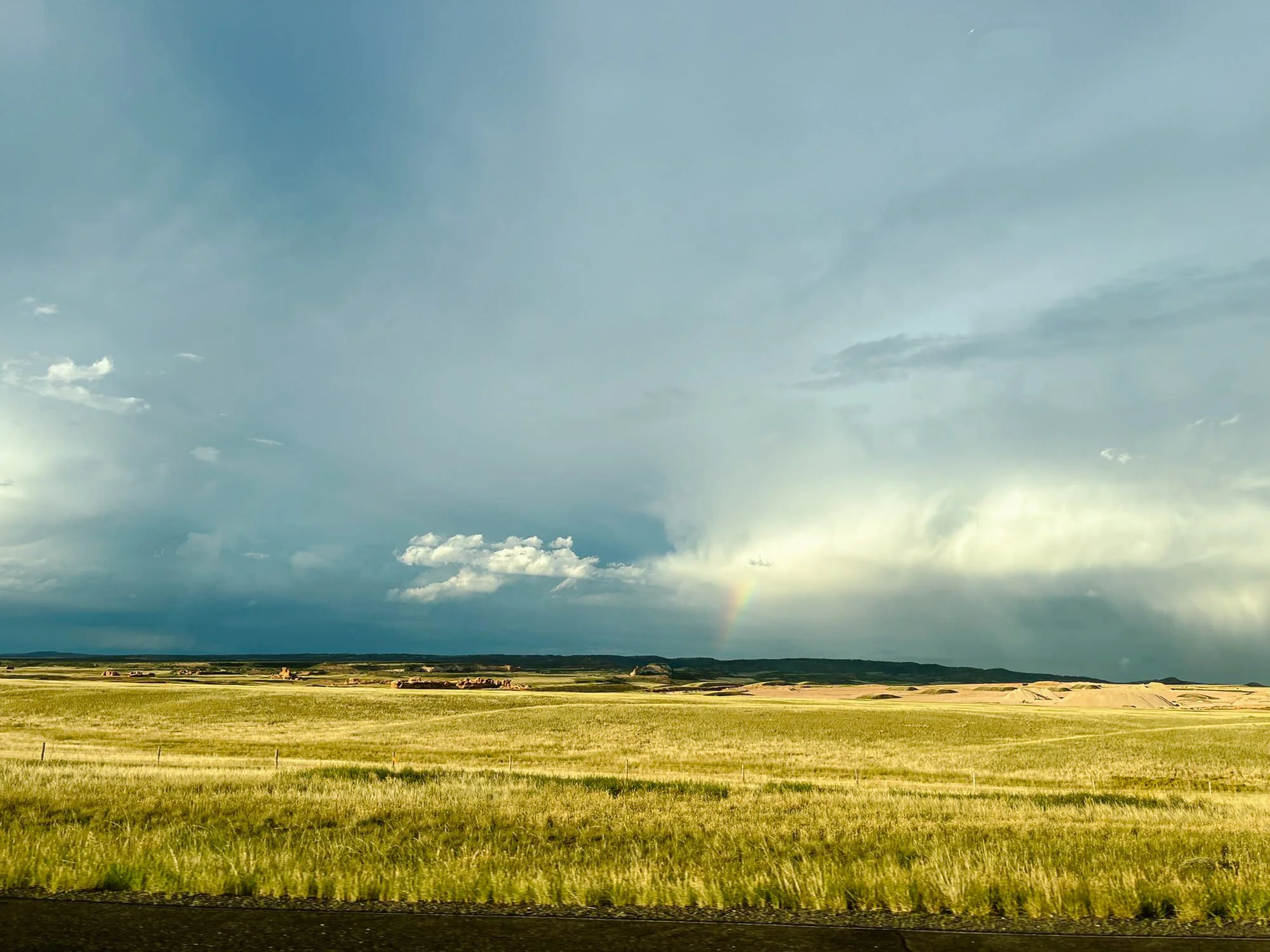 A vast open field with green grass, under a blue sky with scattered clouds and a faint rainbow near the horizon.