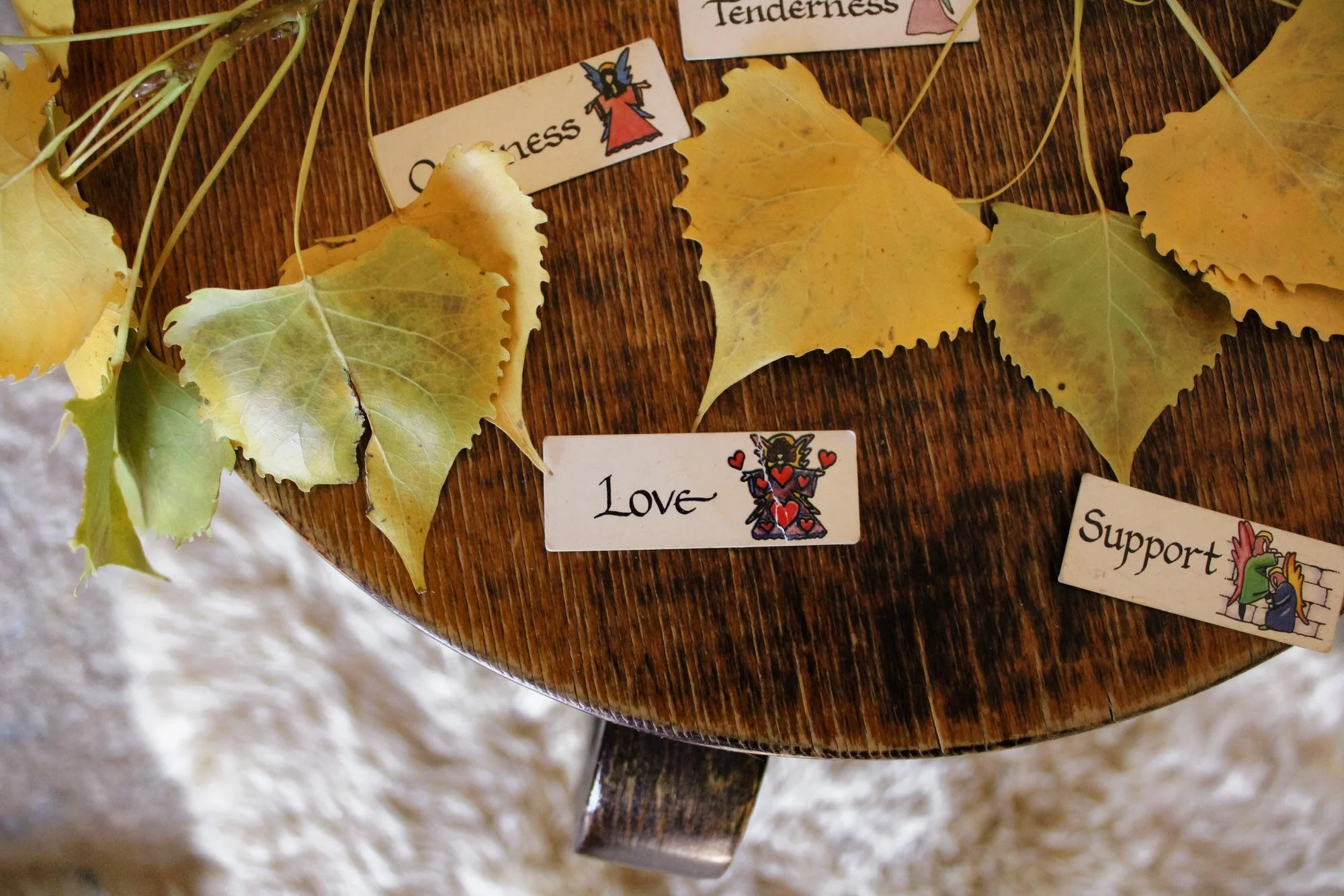 Yellow autumn leaves laid on a wooden surface with small cards featuring words like 'Tenderness', 'Love', and 'Support' along with illustrated images.