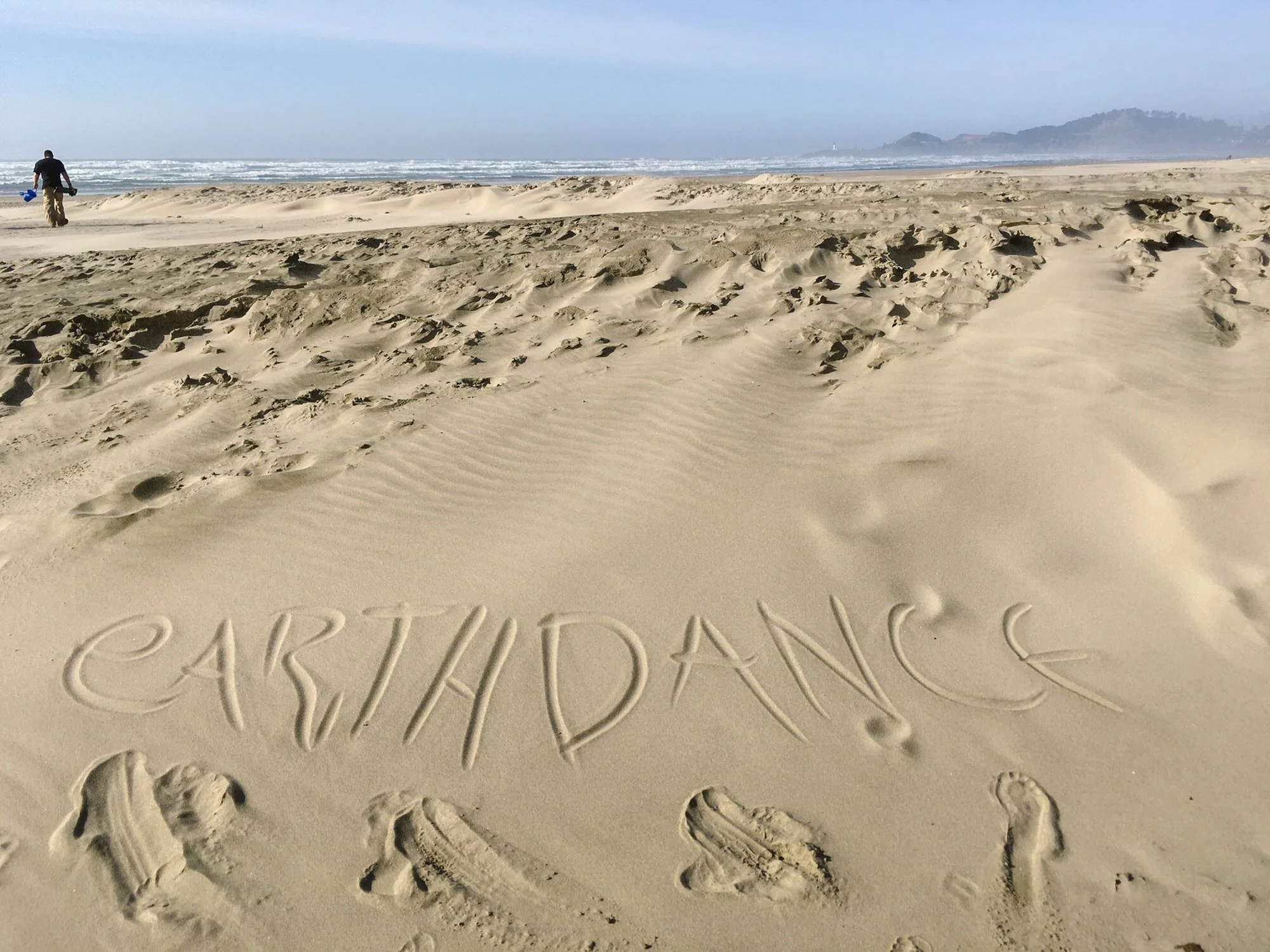 Sand on the beach with the words 'PARTISAN' and 'ASKED' written in large letters in the sand. A man holds a bucket and spade walking near the shoreline. The ocean and a cloudy sky are visible in the background.