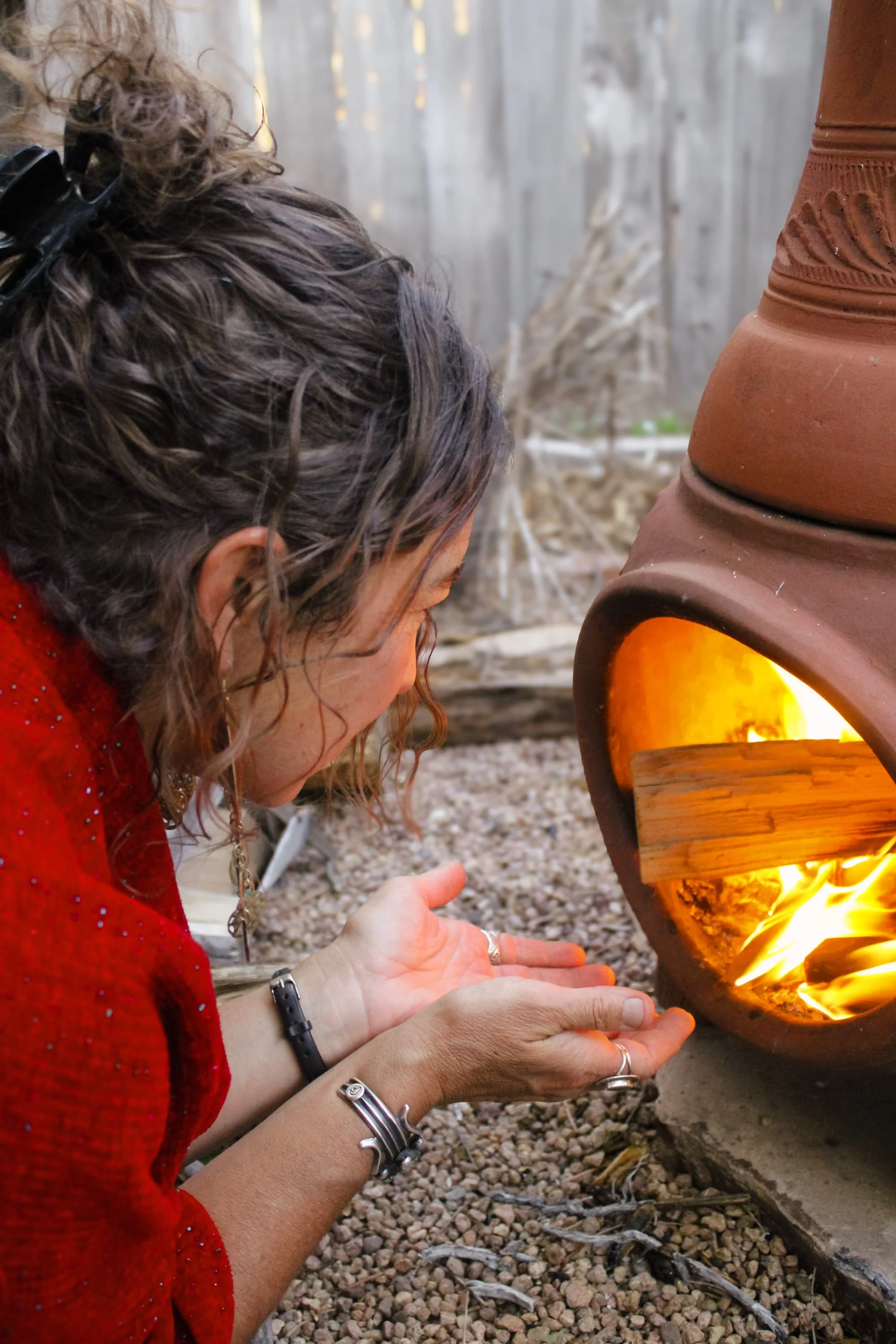 A woman with curly brown hair tied back with a black clip, wearing a red sweater, holds her hands open near a small outdoor fireplace. The fireplace is terracotta-colored with a decorative pattern and glowing inside, with flames visible. The ground is covered in small rocks and gravel, and a wooden fence is in the background.