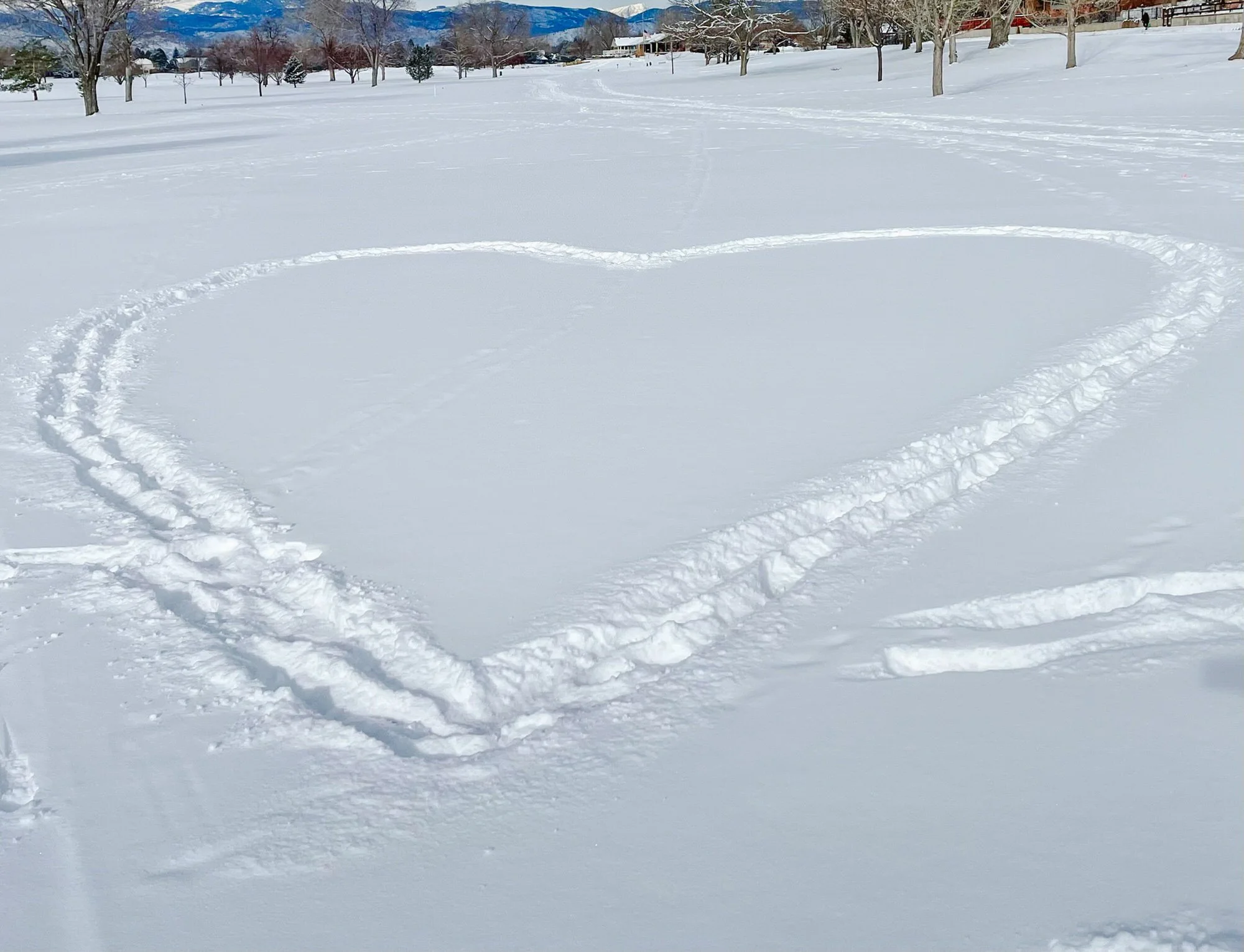 Snowy park with multiple trees, some with bare branches, and distant mountains in the background. A large heart and smaller circle are drawn in the snow, with footprints leading to them.