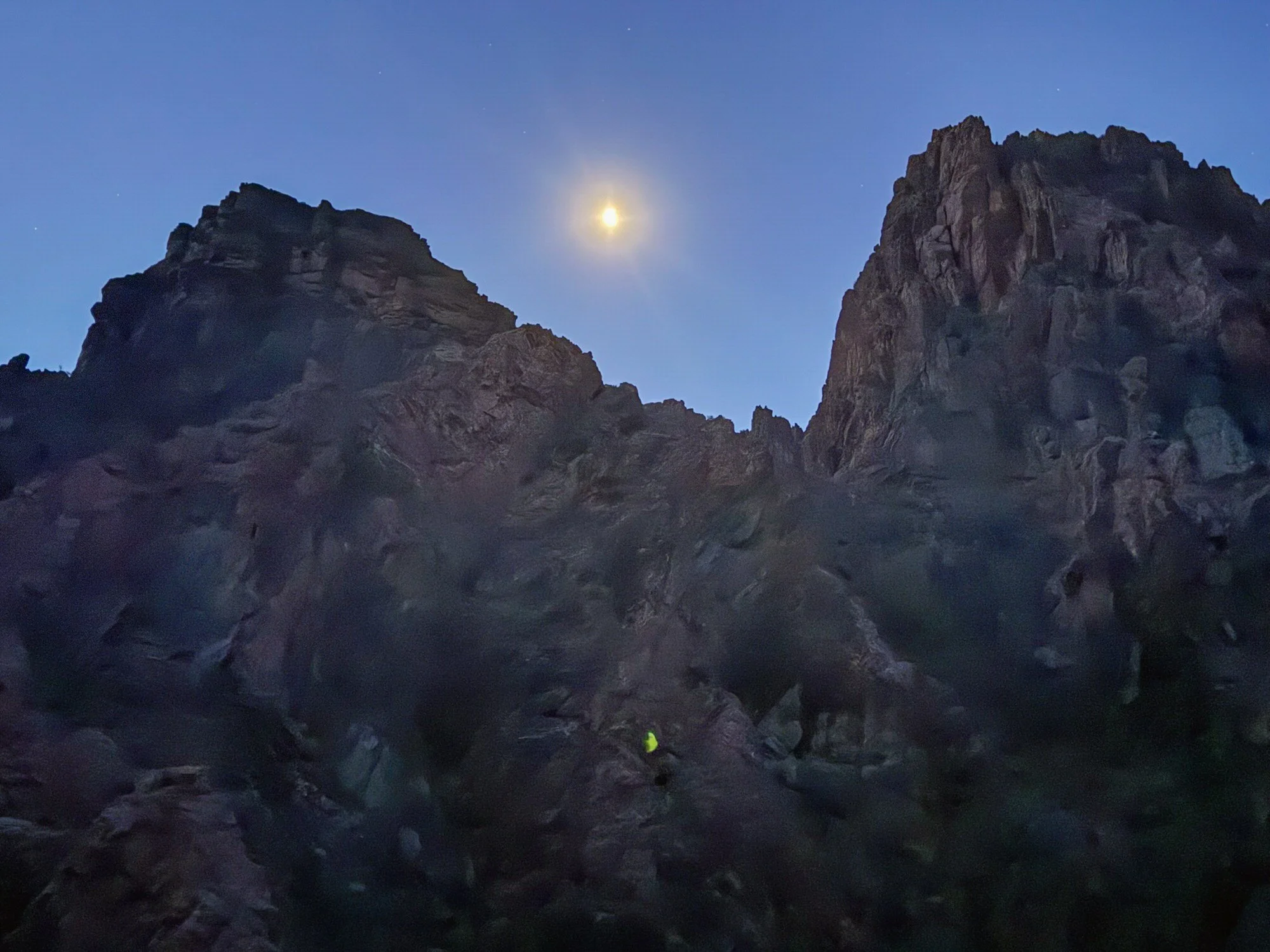 Nighttime scene of a rugged rocky canyon with a bright moon and stars in the sky. A person in a yellow jacket is visible near the bottom of the frame, lost among the large rocks.