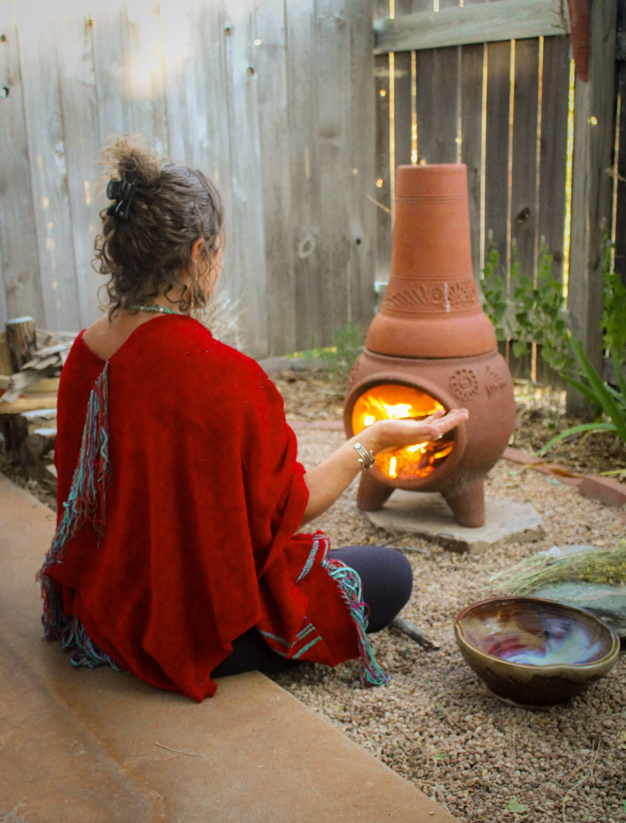 A woman dressed in a red traditional garment kneels by a clay kiln with a fire inside, in an outdoor setting enclosed by a wooden fence. She is engaged in a craft involving the kiln, possibly pottery or ceramic work.
