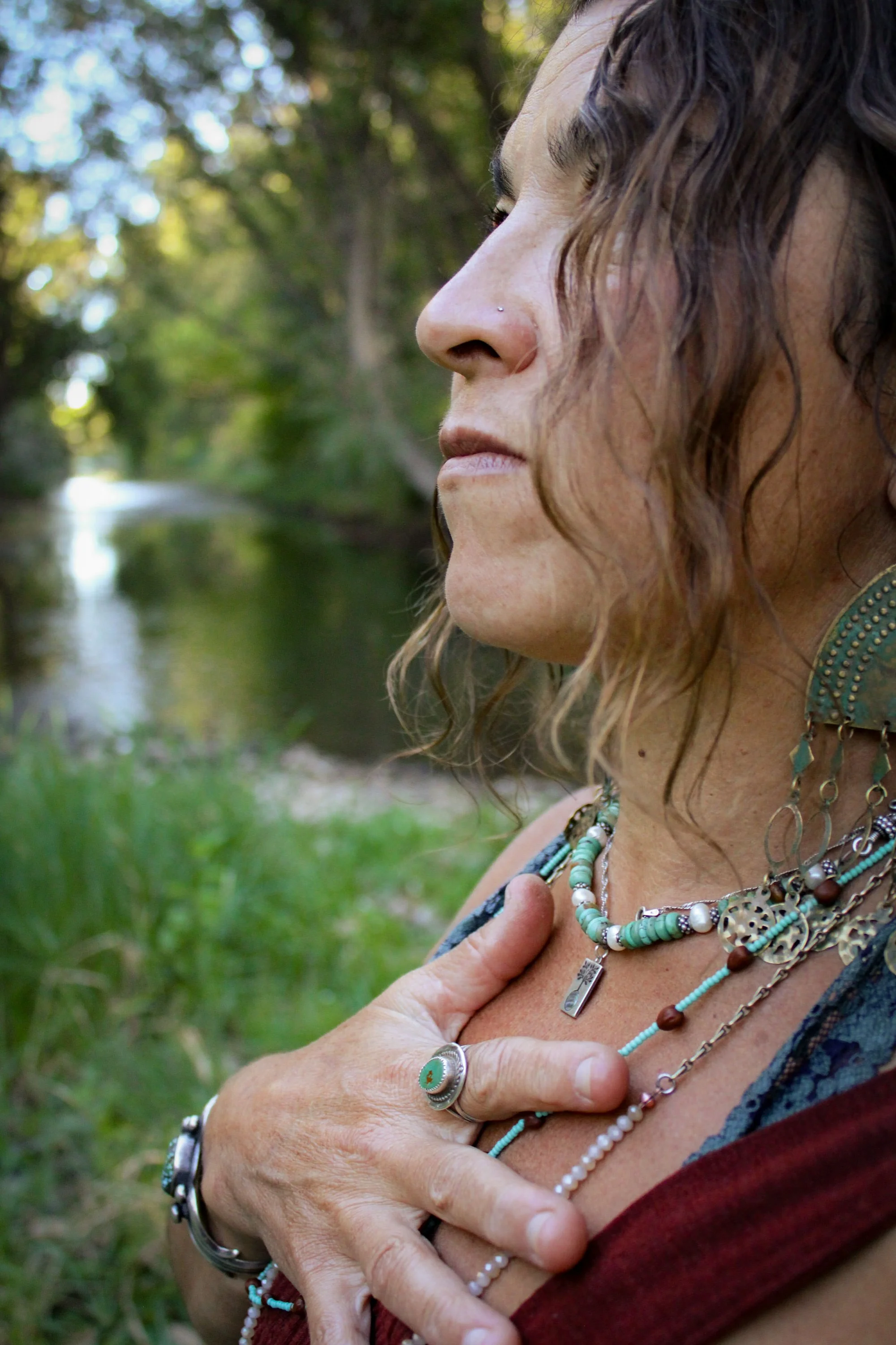 A woman with curly hair and a nose piercing closes her eyes while placing her hand over her chest, wearing multiple necklaces and bracelets in a natural outdoor setting with trees and greenery.