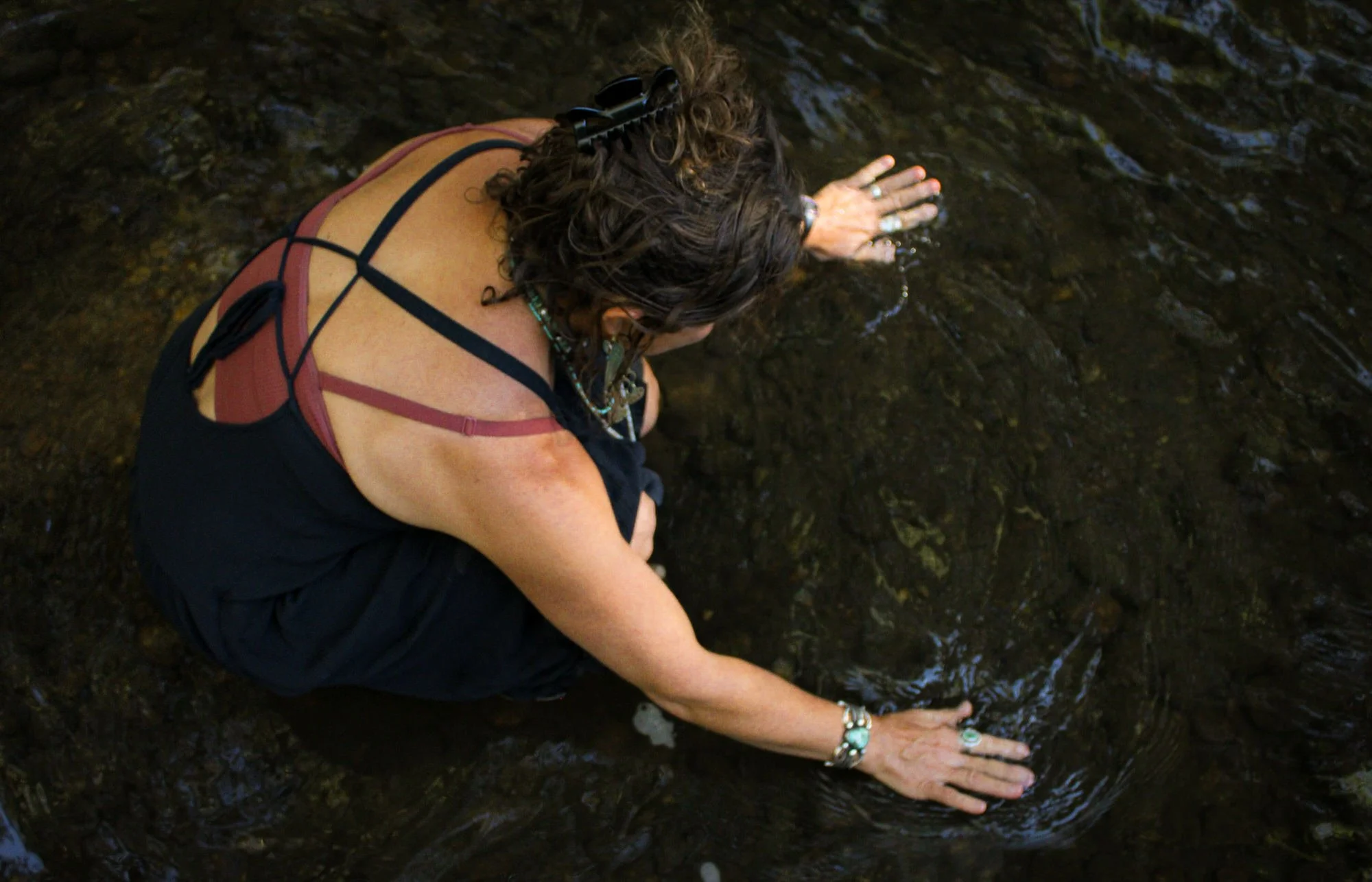 A person with curly hair, wearing colorful jewelry, is kneeling by a stream and touching the water with their hands.
