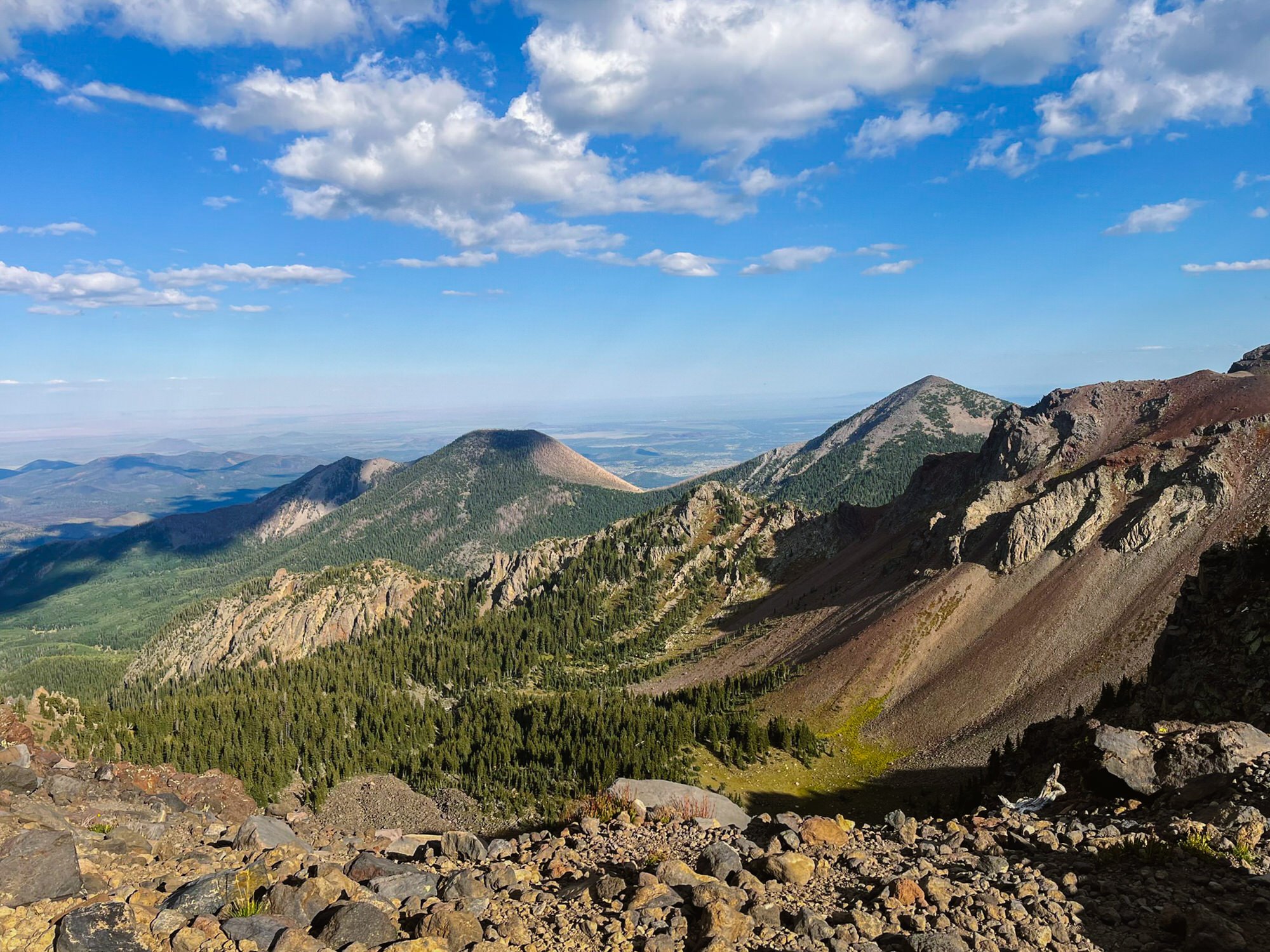 Mountain landscape with rocky foreground, dense green forest on the slopes, and distant mountain peaks under a blue sky with scattered clouds.