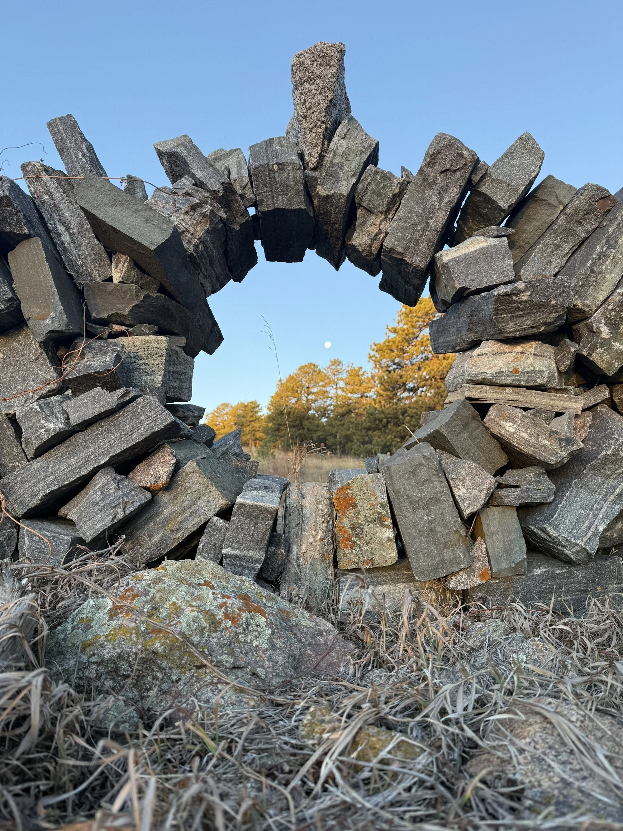 A stone arch made of stacked rocks with a small moon visible in the sky above trees in the background. The foreground features dry grass and moss-covered rocks.