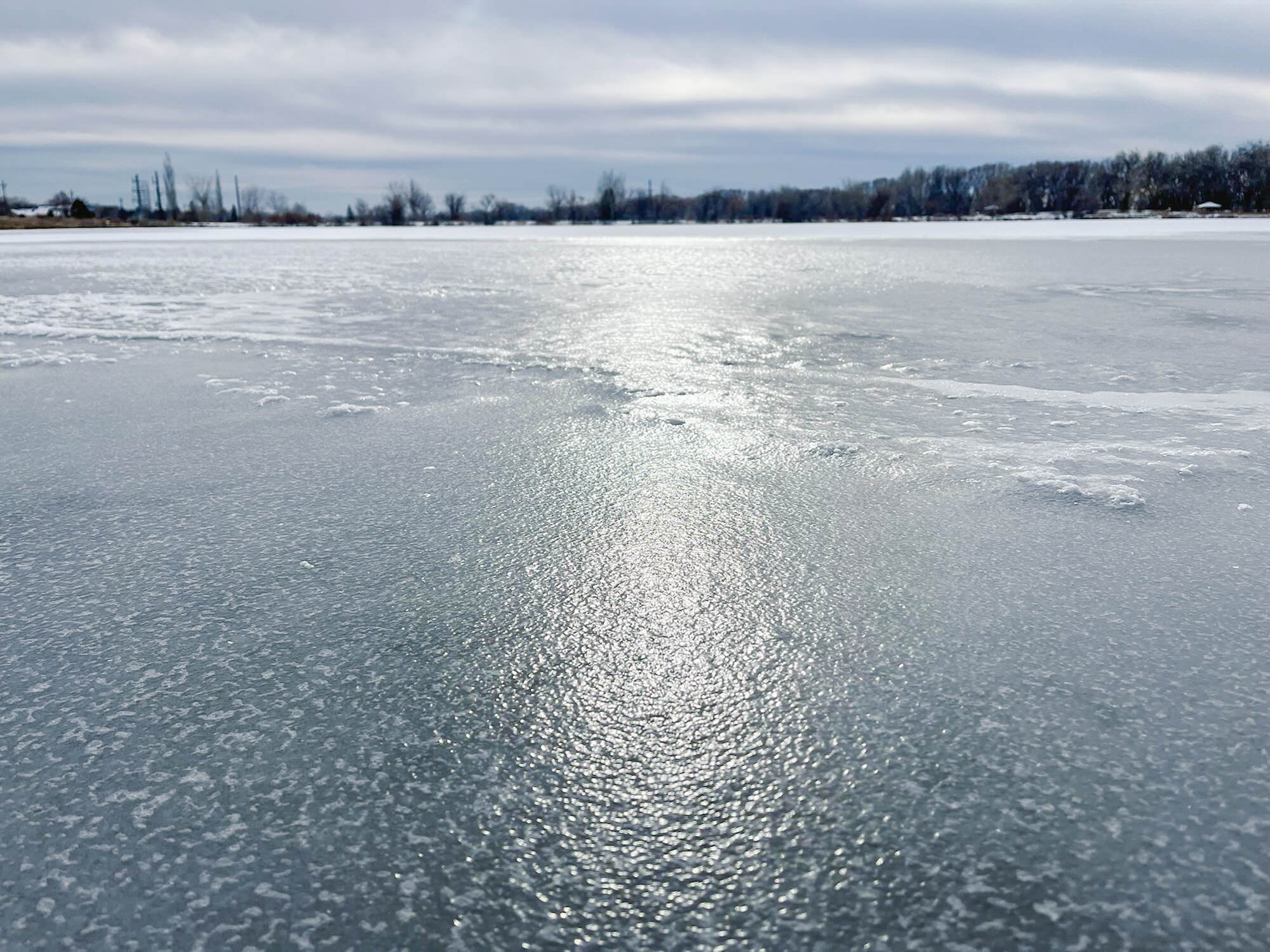 Frozen lake with snow-covered ice surface and trees and buildings in the distance under a cloudy sky.