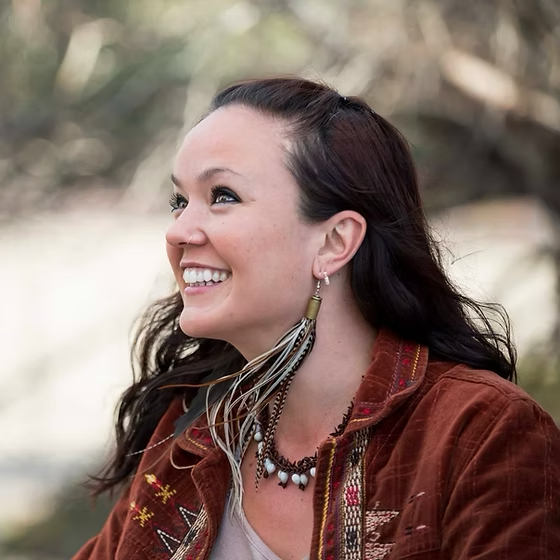 A woman with dark hair smiling outdoors, wearing a brown jacket with colorful embroidery, layered necklaces, and feather earrings.