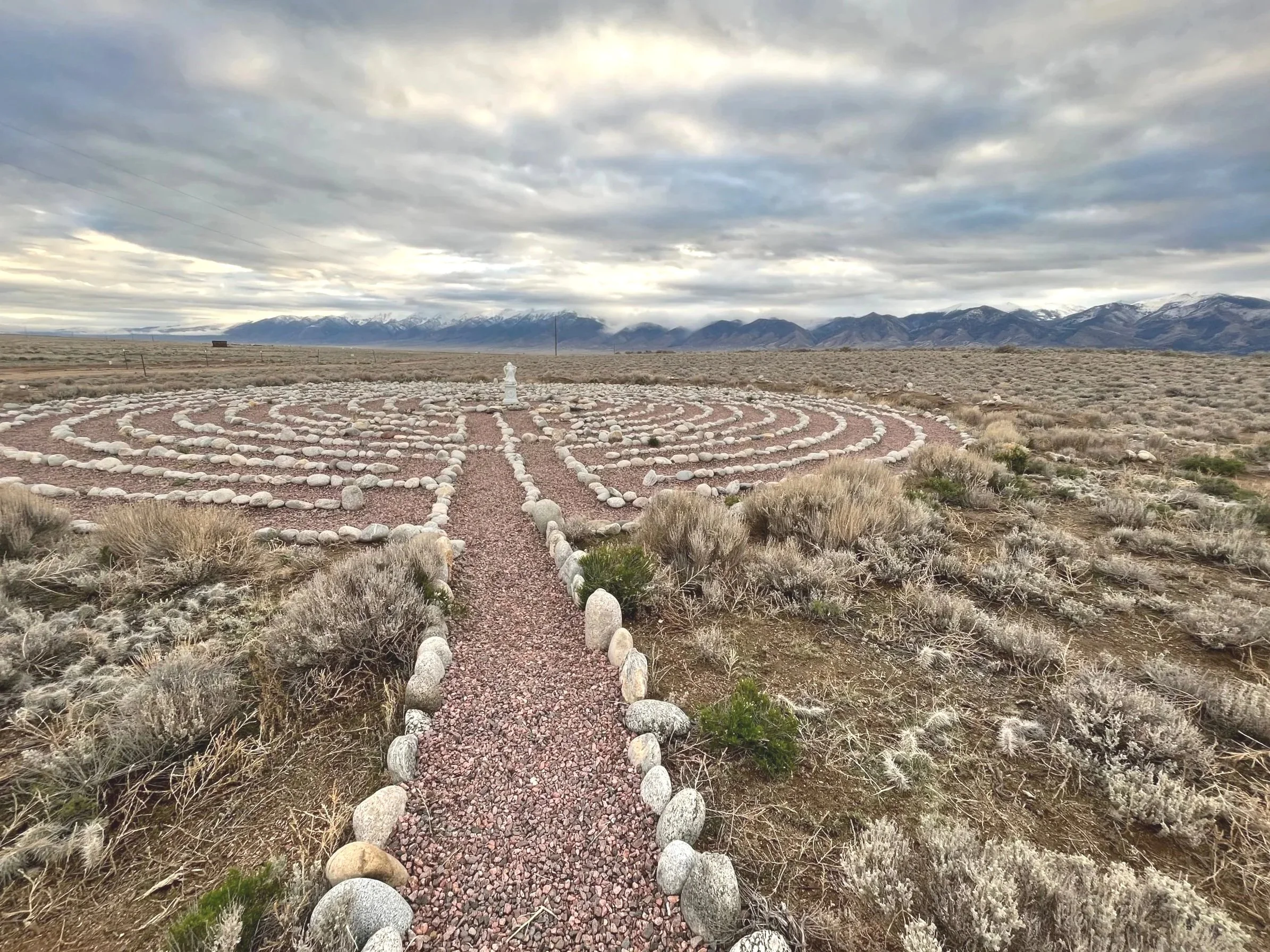 A landscaped garden with a circular rock pattern, surrounded by desert plants, with mountains and cloudy sky in the background.