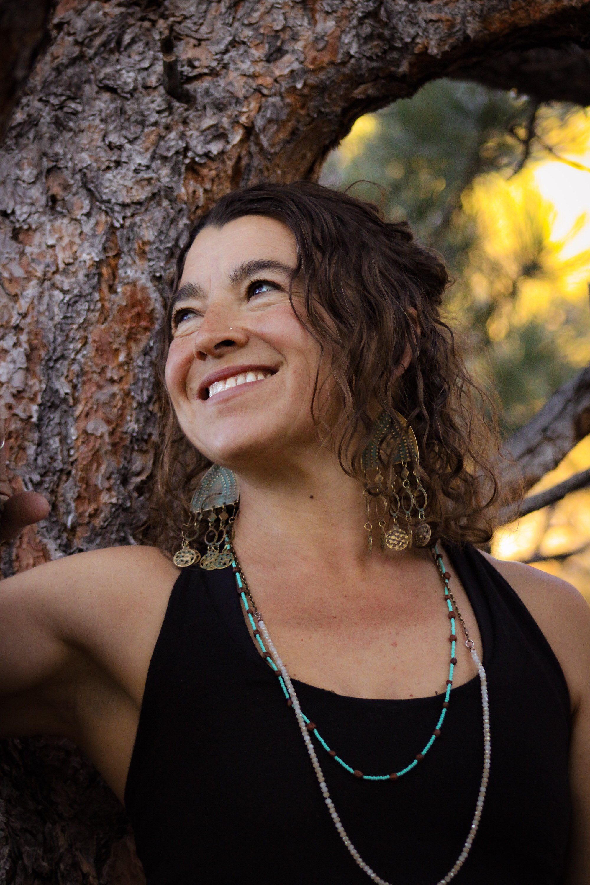 A woman with curly brown hair smiling outdoors by a tree during sunset, wearing earrings and layered necklaces.