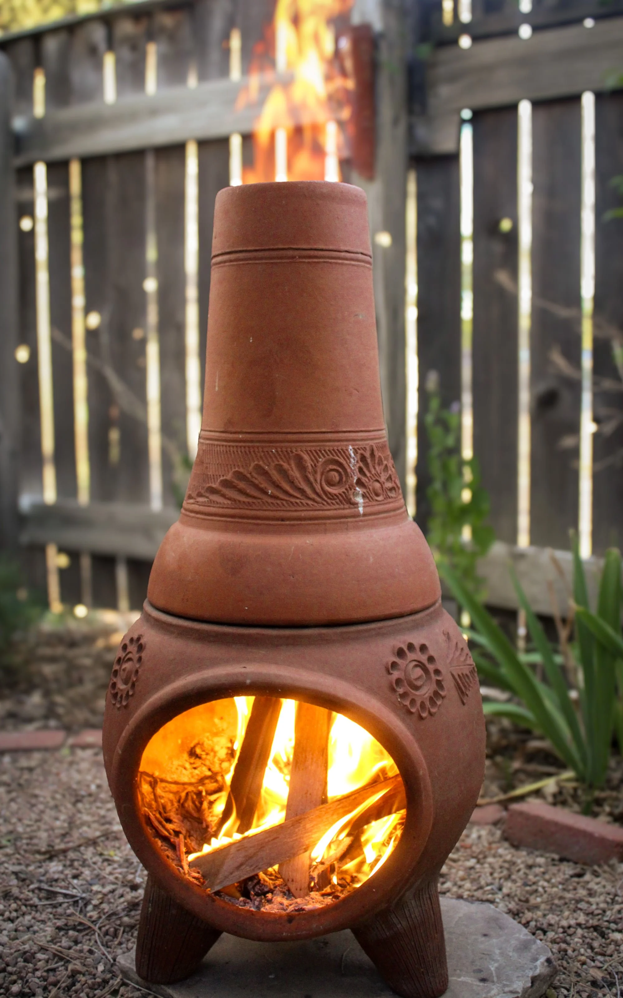 Terracotta wind chime with a small fire inside, set outdoors with a wooden fence and green plants in the background.