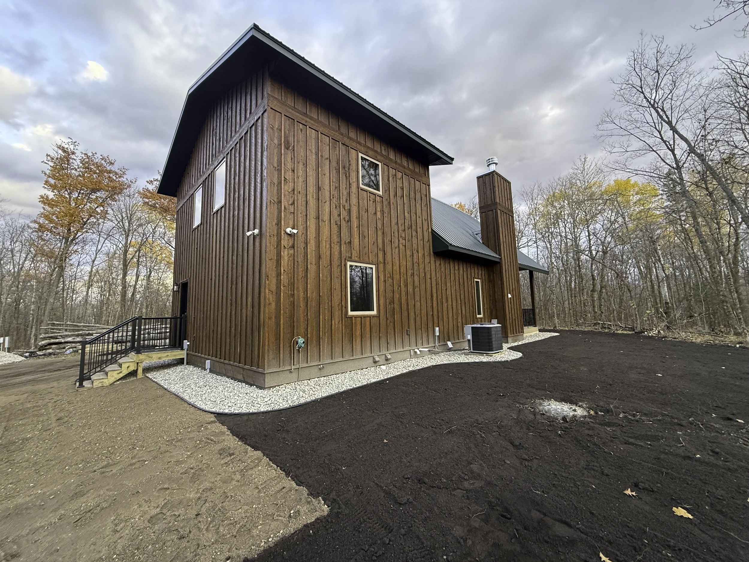 A modern two-story house with wood siding and a metal roof, surrounded by trees with fall foliage, under a cloudy sky.