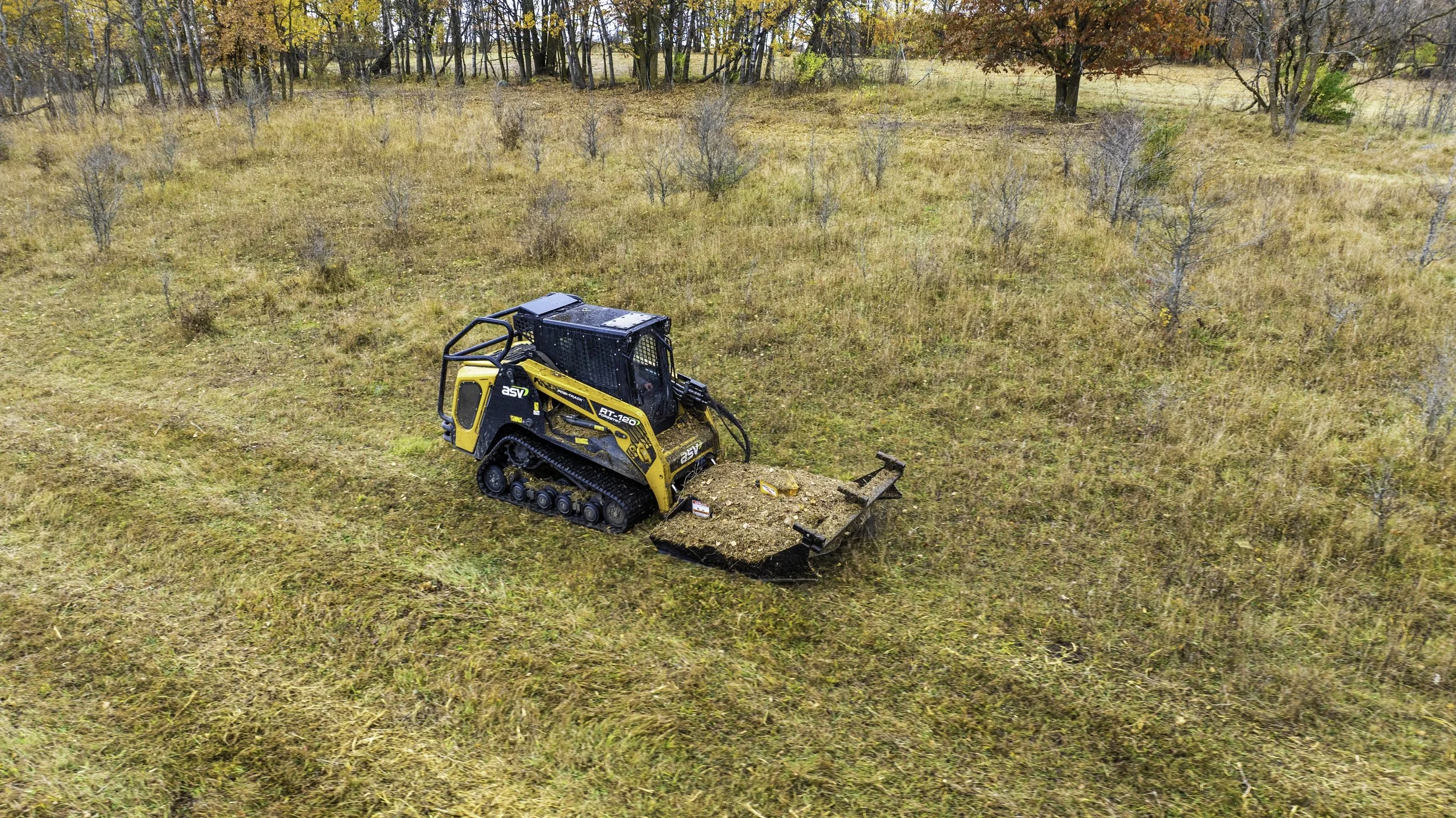 A small yellow and black tracked bulldozer moving soil across a grassy field with sparse trees and autumn foliage in the background.