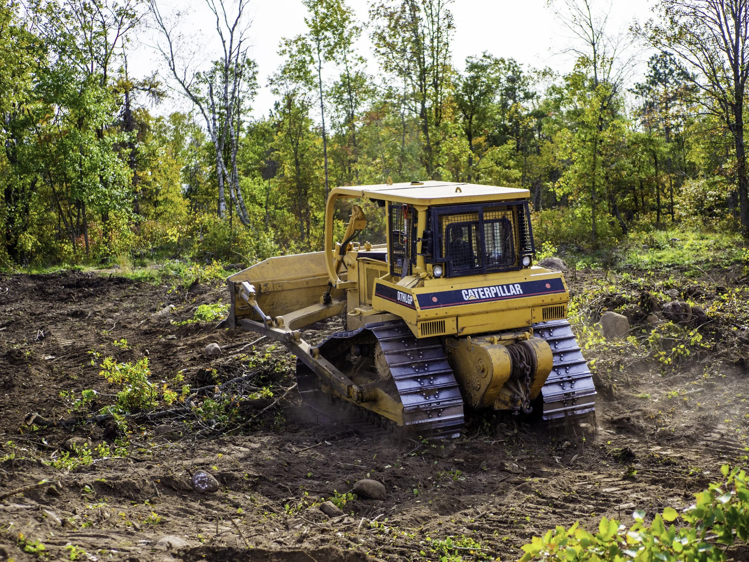 A yellow Caterpillar bulldozer moving soil and vegetation in a forested area during daytime.