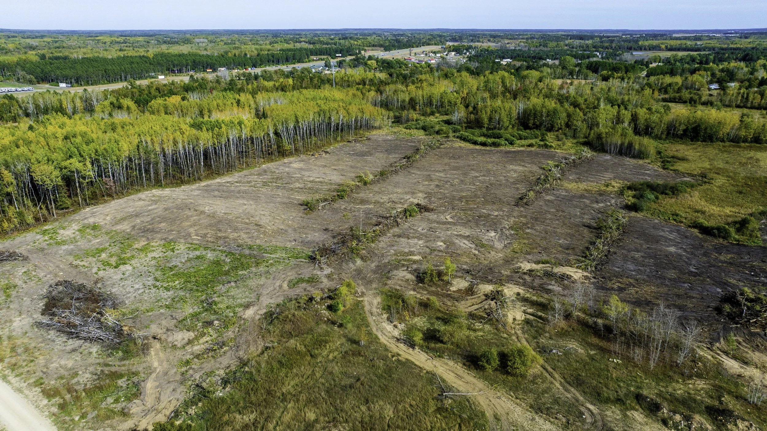 Aerial view of land clearing showing patches of disturbed soil, some tree debris, and partially cleared areas next to a forest with green and yellow foliage, with a highway and trees in the background.