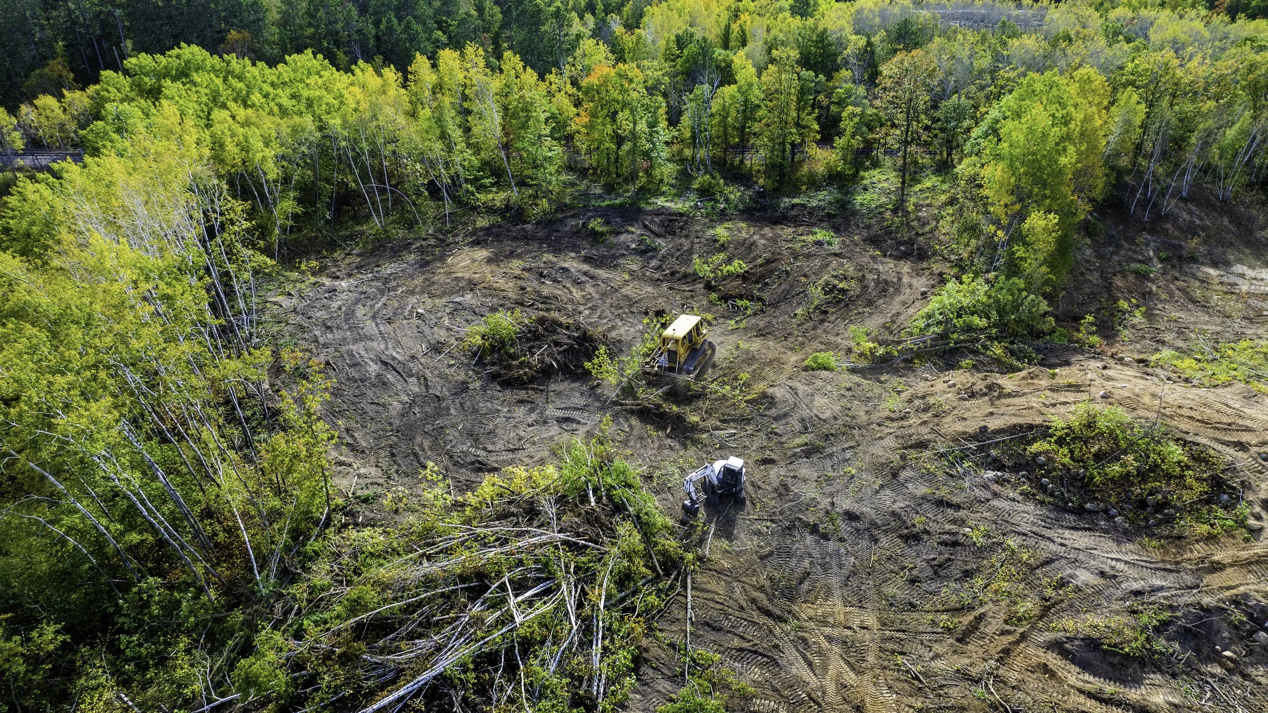 Aerial view of a forest clearing with a bulldozer and an excavator working on land clearing, surrounded by trees in spring foliage.